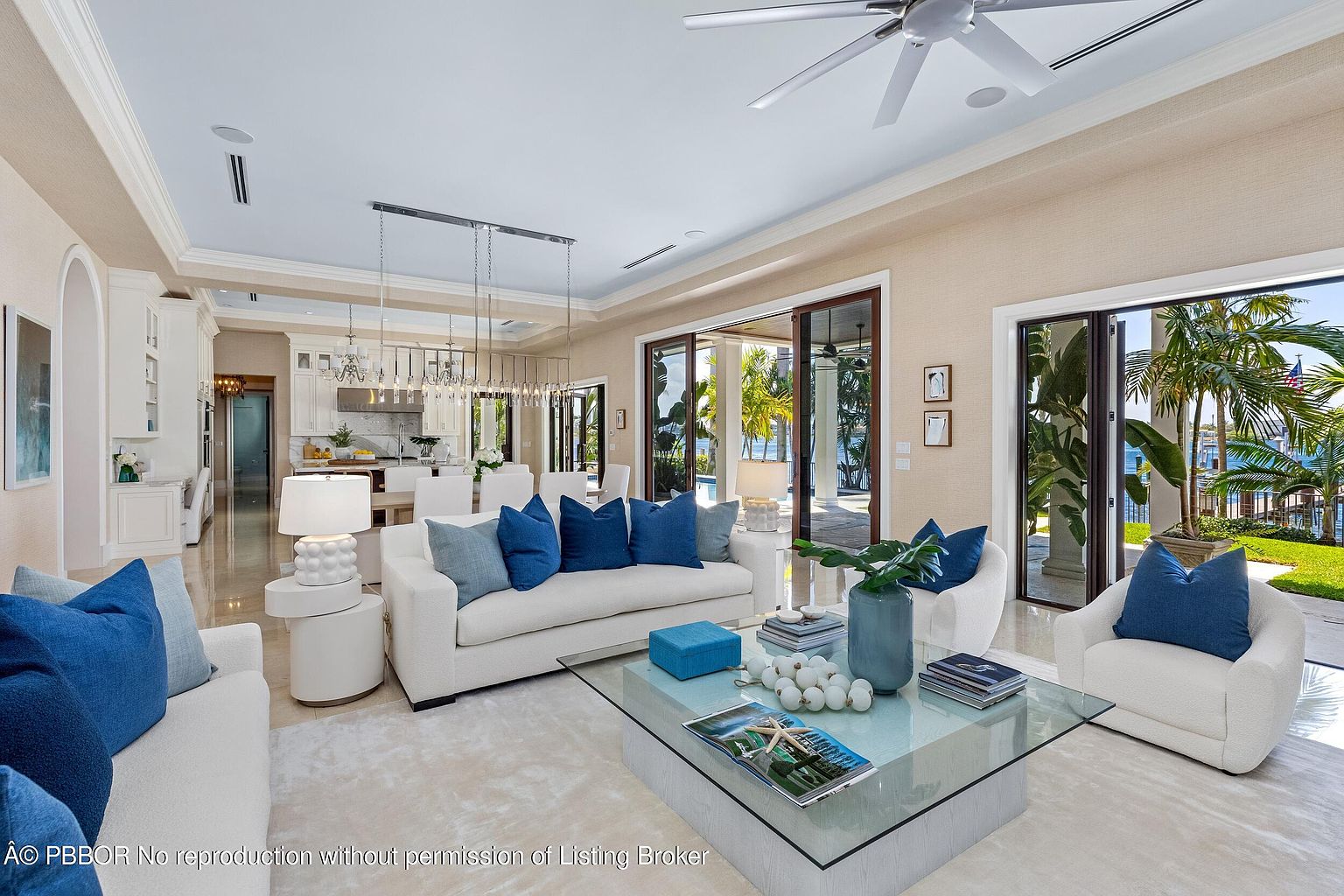 This is a bright and airy living room featuring a neutral color palette with pops of blue from the throw pillows. The room is furnished with white sofas and armchairs, a glass-topped coffee table, and a light-colored rug. Large sliding glass doors provide access to an outdoor area and offer views of the water, creating a seamless indoor-outdoor living experience.