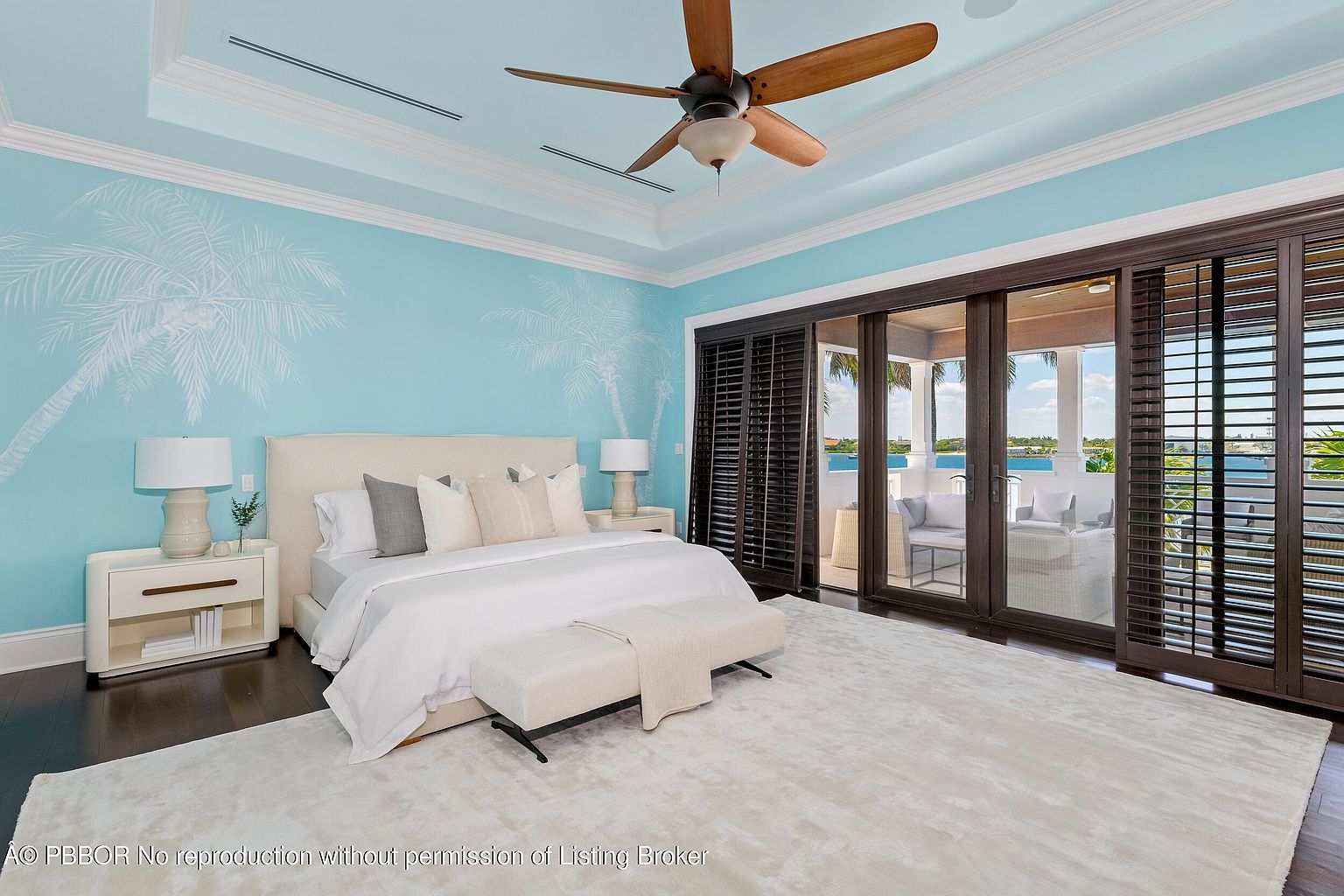 This is a bright and airy primary bedroom featuring a king-size bed with white linens and decorative pillows. The room has a light blue accent wall with palm tree designs, complemented by dark wood flooring and a large white area rug. Sliding glass doors with dark wood frames lead to an outdoor balcony, enhancing the room's spacious feel and providing natural light.