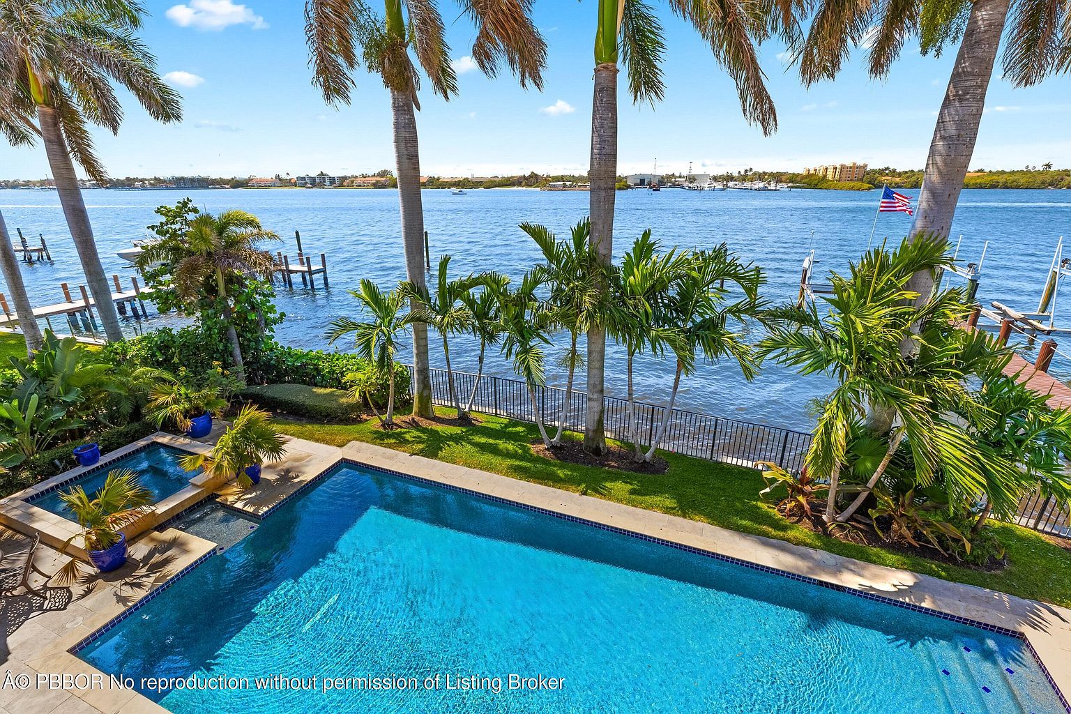 This is an aerial view of a luxurious backyard featuring a sparkling blue pool and spa area, surrounded by lush tropical landscaping including palm trees. The property backs onto a scenic waterfront with a view of the water, docks, and distant buildings, enhancing the sense of privacy and exclusivity. An American flag is visible on one of the docks.