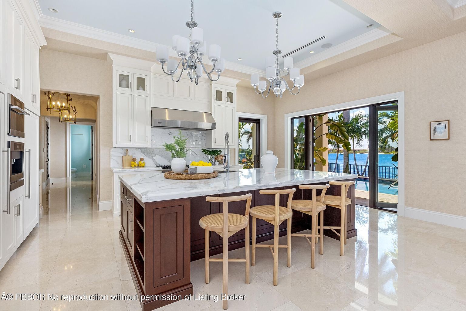 This is a bright and luxurious kitchen featuring a large center island with seating, marble countertops, and custom cabinetry. The kitchen is well-lit by chandeliers and natural light streaming in from the sliding glass doors that offer a view of the pool and waterfront. The overall impression is one of elegance and high-end design.