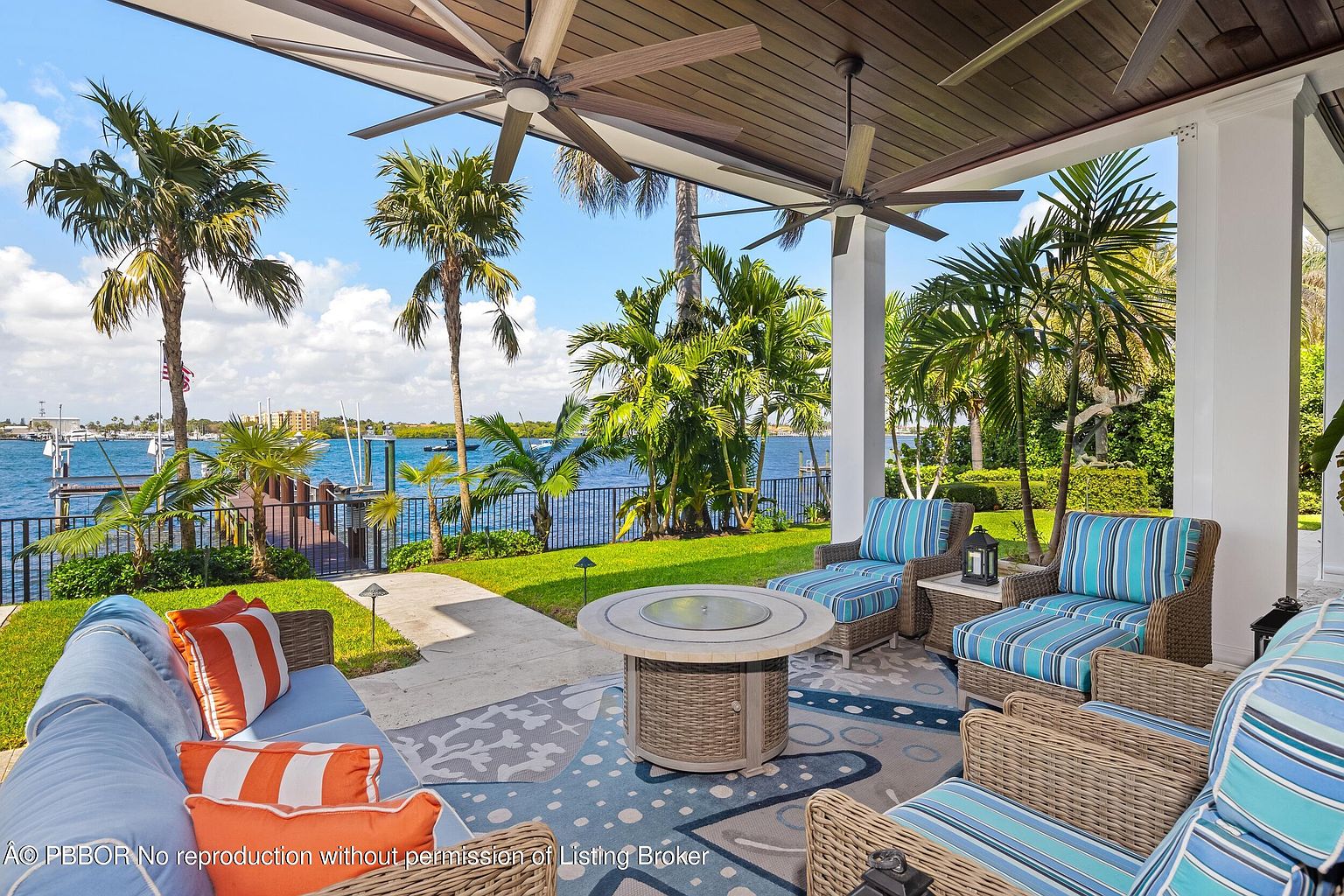 This image showcases a luxurious outdoor patio area with comfortable wicker furniture, including sofas and armchairs with blue and white striped cushions and orange pillows. A round fire pit table sits in the center of a patterned rug, creating a cozy gathering space. The patio overlooks a waterfront view with palm trees and a dock, enhancing the property's appeal for outdoor living and entertaining.
