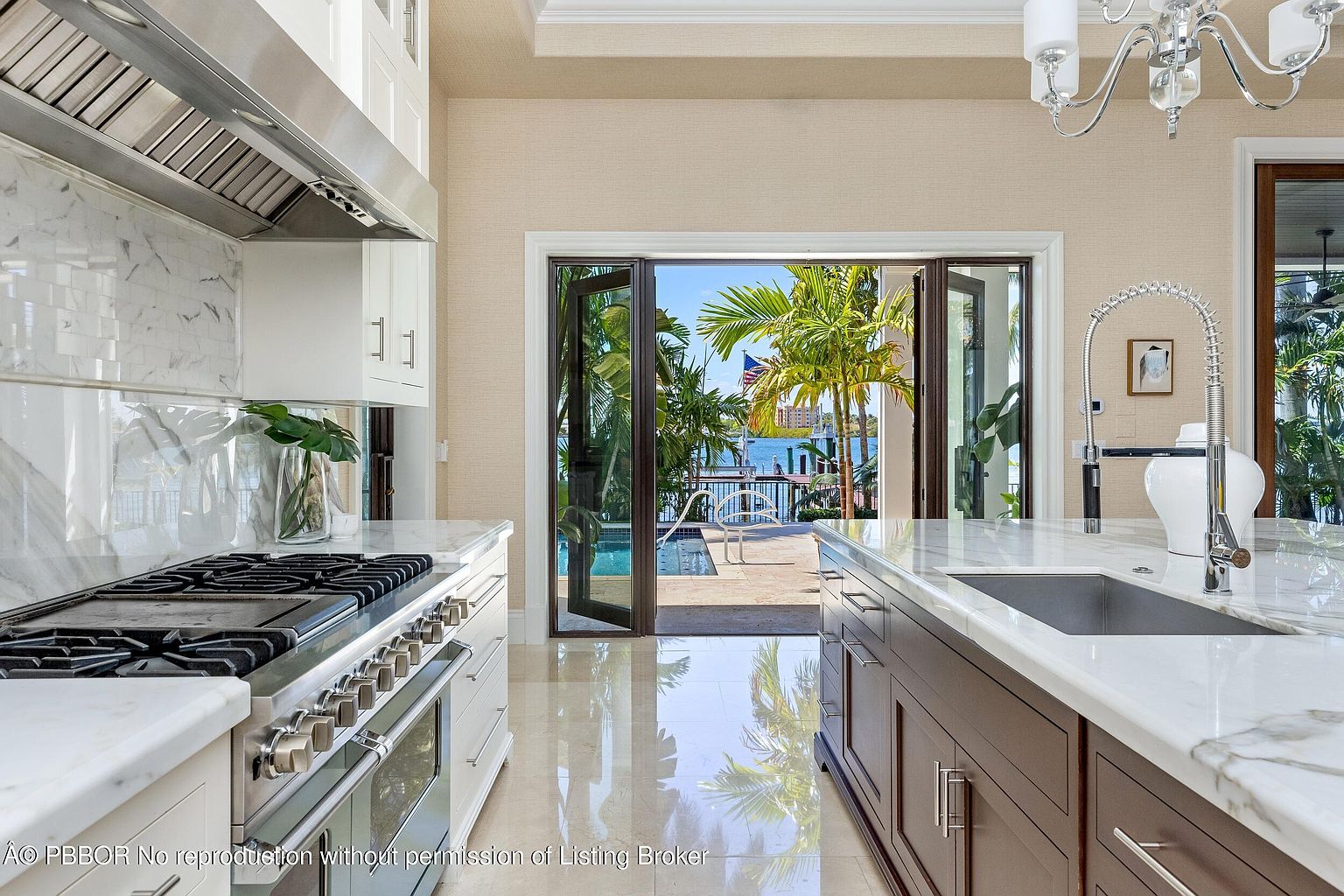 This is a bright and modern kitchen featuring a professional-grade gas range with a stainless steel hood, white cabinetry, and a large island with a marble countertop. The kitchen opens to an outdoor patio and pool area through folding glass doors, providing a seamless indoor-outdoor living experience. The flooring is a polished tile, reflecting the natural light and enhancing the spacious feel of the room.