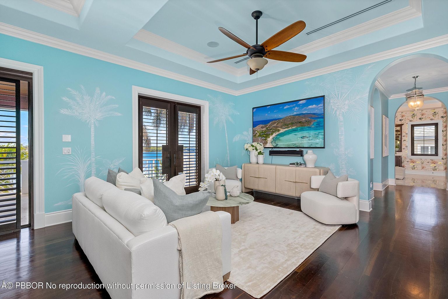 This is an interior shot of a living room featuring light blue walls adorned with subtle palm tree designs. The room is furnished with a white sofa, a round coffee table, and a light-colored media console beneath a large television displaying a coastal scene. The dark wood flooring and ceiling fan add a touch of warmth to the space.