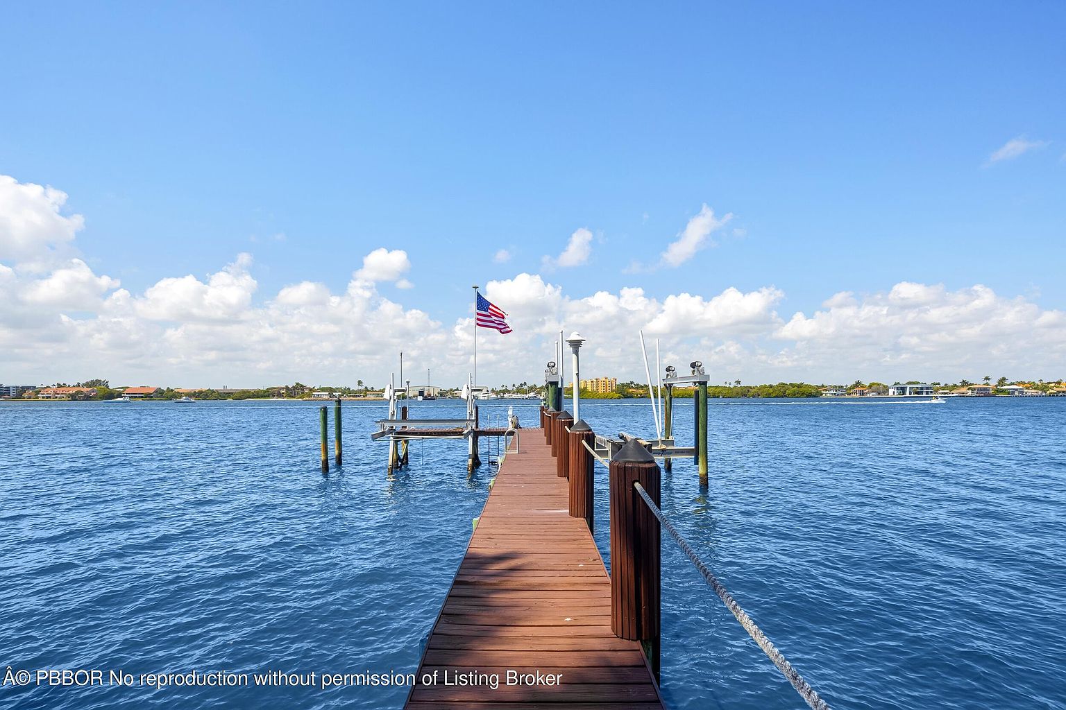 This image showcases a wooden pier extending into a serene body of water, likely a bay or ocean inlet. An American flag waves gently at the end of the pier, adding a patriotic touch. The scene is bathed in natural light under a partly cloudy sky, creating a peaceful and inviting atmosphere.