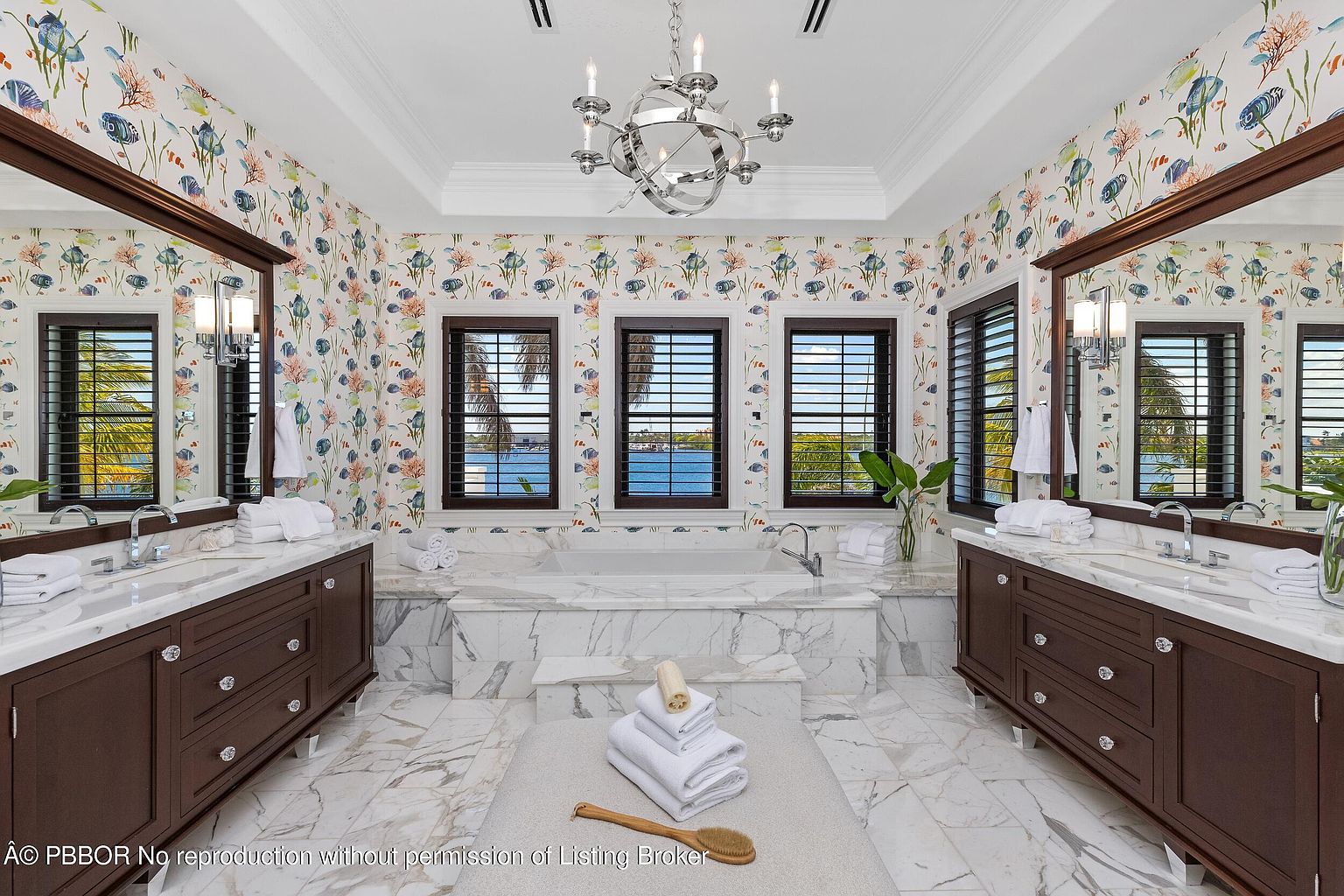 This is a luxurious primary bathroom featuring dual vanities with dark wood cabinetry and marble countertops. The walls are adorned with a whimsical fish-themed wallpaper, and natural light floods the space through three windows overlooking a waterfront view. A freestanding tub is nestled between the vanities, creating a spa-like atmosphere.