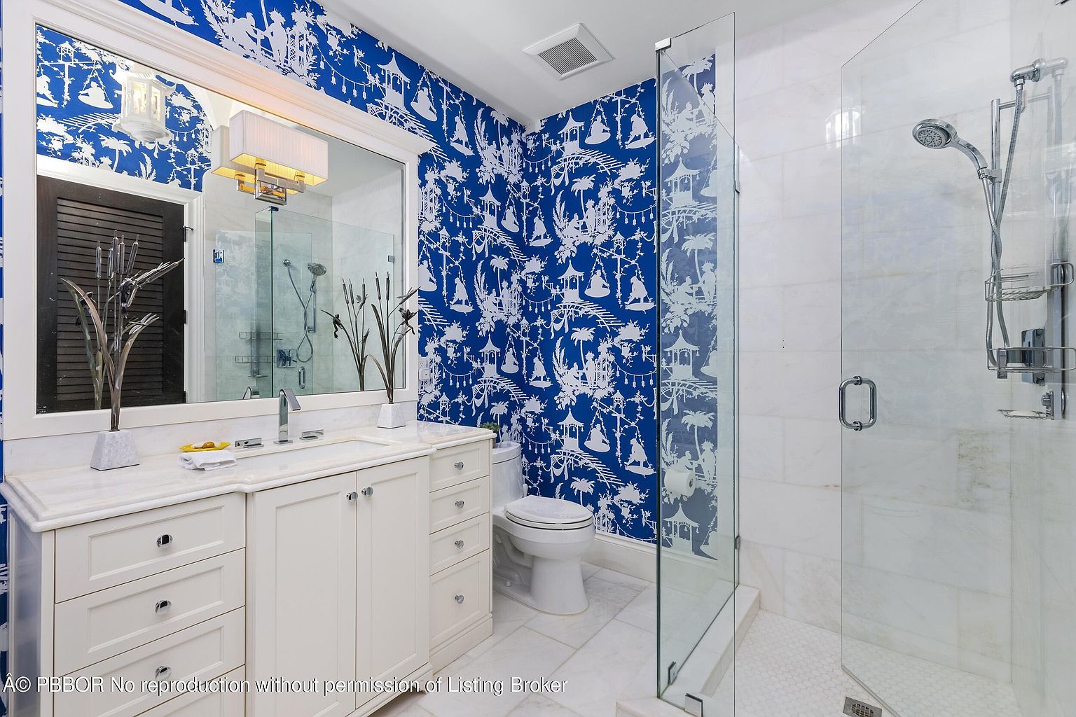 This is a well-lit bathroom featuring a white vanity with marble countertop, a toilet, and a glass-enclosed shower. The walls are adorned with a blue and white patterned wallpaper, adding a unique and stylish touch to the space. The overall impression is clean, bright, and tastefully decorated.