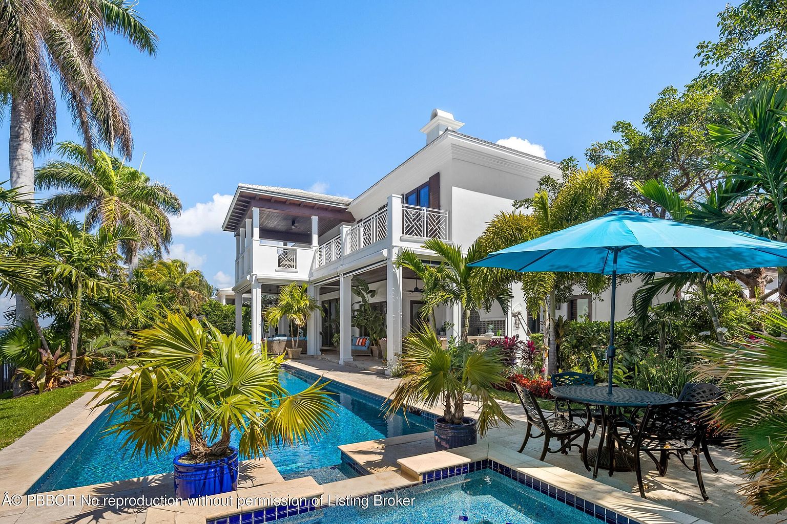 This image showcases a luxurious backyard pool area with a two-story white house in the background. The pool features a clear blue water and is surrounded by lush tropical landscaping, including palm trees and potted plants. An outdoor dining set with a blue umbrella provides a relaxing space next to the pool, creating an inviting and upscale atmosphere.