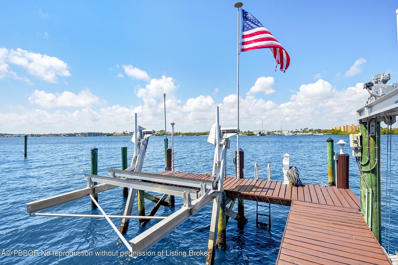 This image showcases a waterfront deck featuring a boat lift and an American flag waving in the breeze. The deck is constructed of wood and provides direct access to the water. The scene is set against a backdrop of a clear blue sky with scattered clouds, creating a serene and inviting atmosphere.