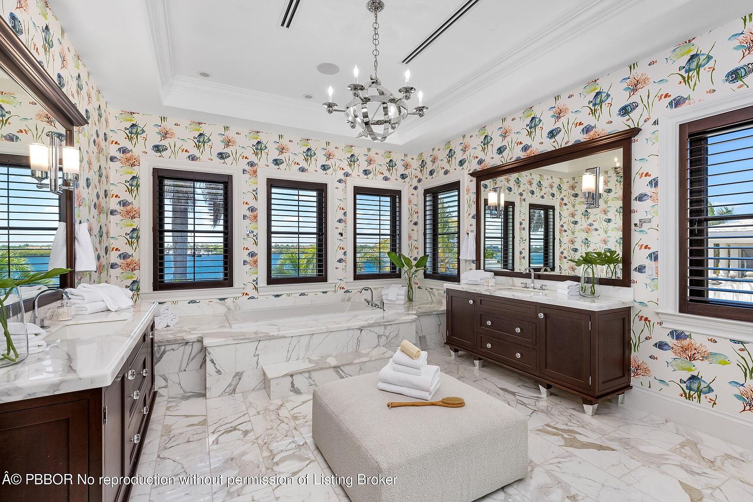 This is a luxurious primary bathroom featuring marble flooring and walls with a unique fish-themed wallpaper. The room includes a large soaking tub, dual vanities with dark wood cabinetry, and a central ottoman. Natural light floods the space through multiple windows with dark shutters, enhancing the serene and upscale atmosphere.