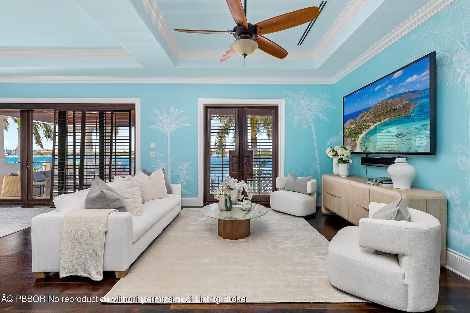 This is an interior shot of a living room featuring a light blue color scheme with palm tree accents. The room is furnished with a white sofa, two white armchairs, and a round coffee table on a light-colored rug. A large television is mounted on the wall, and natural light streams in through the sliding glass doors, offering a view of the ocean.