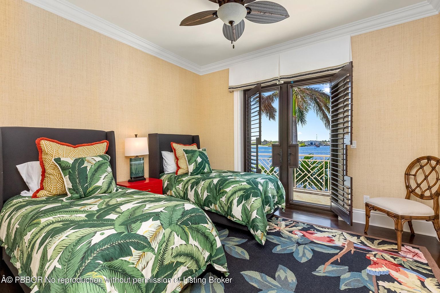 This is a guest bedroom featuring two twin beds with tropical-themed bedding, dark headboards, and red side tables. The room has a light yellow textured wallpaper and a ceiling fan. French doors with shutters lead to a balcony with a view of the water, and a patterned rug adds a touch of color to the dark wood floor. A decorative chair sits to the right of the doors.
