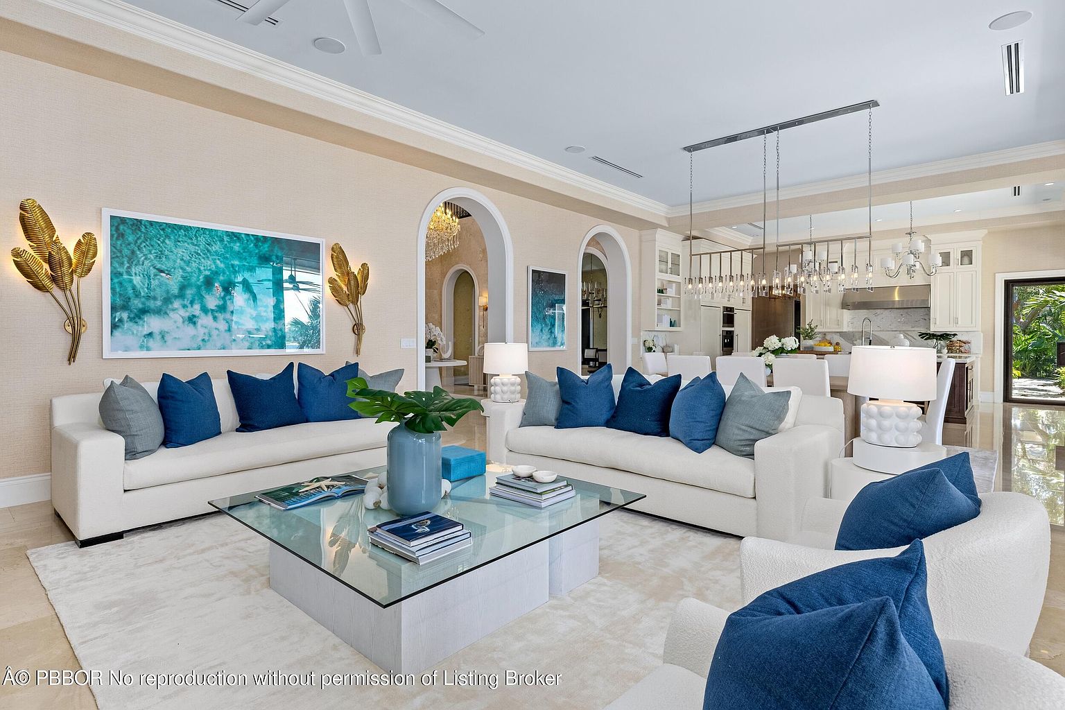 This is an interior shot of a luxurious living room featuring two white sofas adorned with blue pillows, a glass-topped coffee table, and a plush white rug. The room is decorated with modern art and sconces, and it opens to a kitchen area through arched doorways, creating an open and airy feel. The overall impression is one of elegance and comfort.