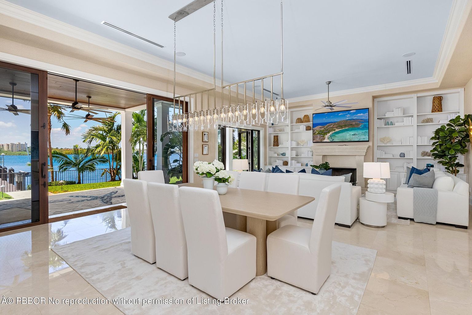 This interior shot showcases a luxurious dining area seamlessly connected to a living space and an outdoor view. The dining area features a light wood table surrounded by white upholstered chairs, illuminated by a modern chandelier. The open floor plan leads to a living room with built-in shelving and a large television, while sliding glass doors offer a view of a waterfront landscape with palm trees.