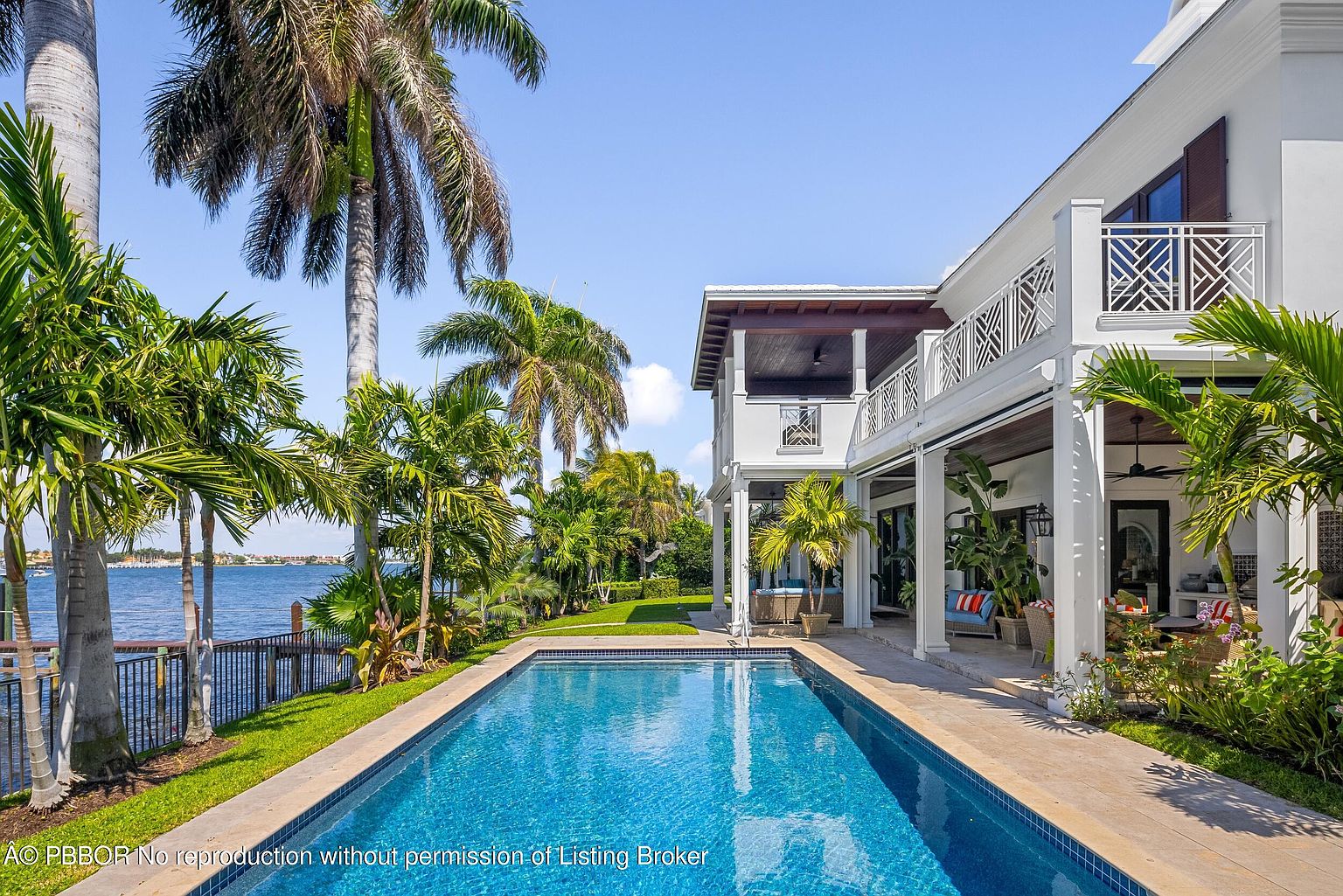 This image showcases a luxurious backyard featuring a long, rectangular swimming pool with clear blue water, surrounded by a tiled patio and lush green lawn. The pool is adjacent to a grand two-story white house with a covered patio and balcony, framed by tropical palm trees and waterfront views, creating an inviting and serene outdoor living space.