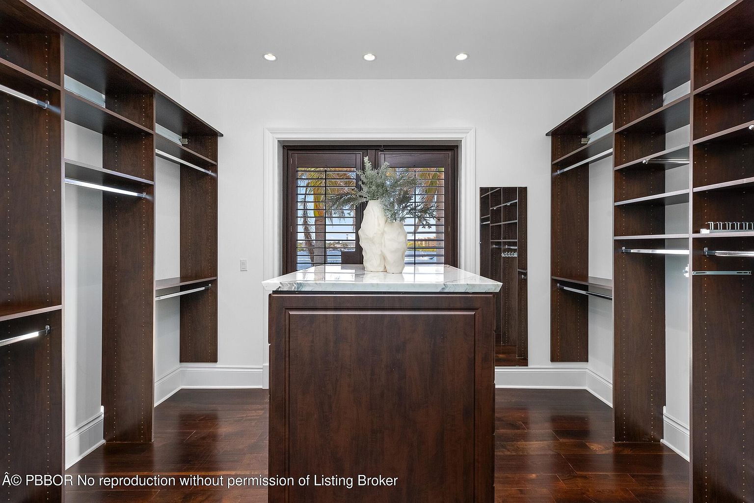 This is a well-organized walk-in closet featuring dark wood shelving and cabinetry on both sides, providing ample storage space. A central island with a marble countertop adds functionality and style. The closet is well-lit and appears spacious, enhancing the appeal for potential buyers.