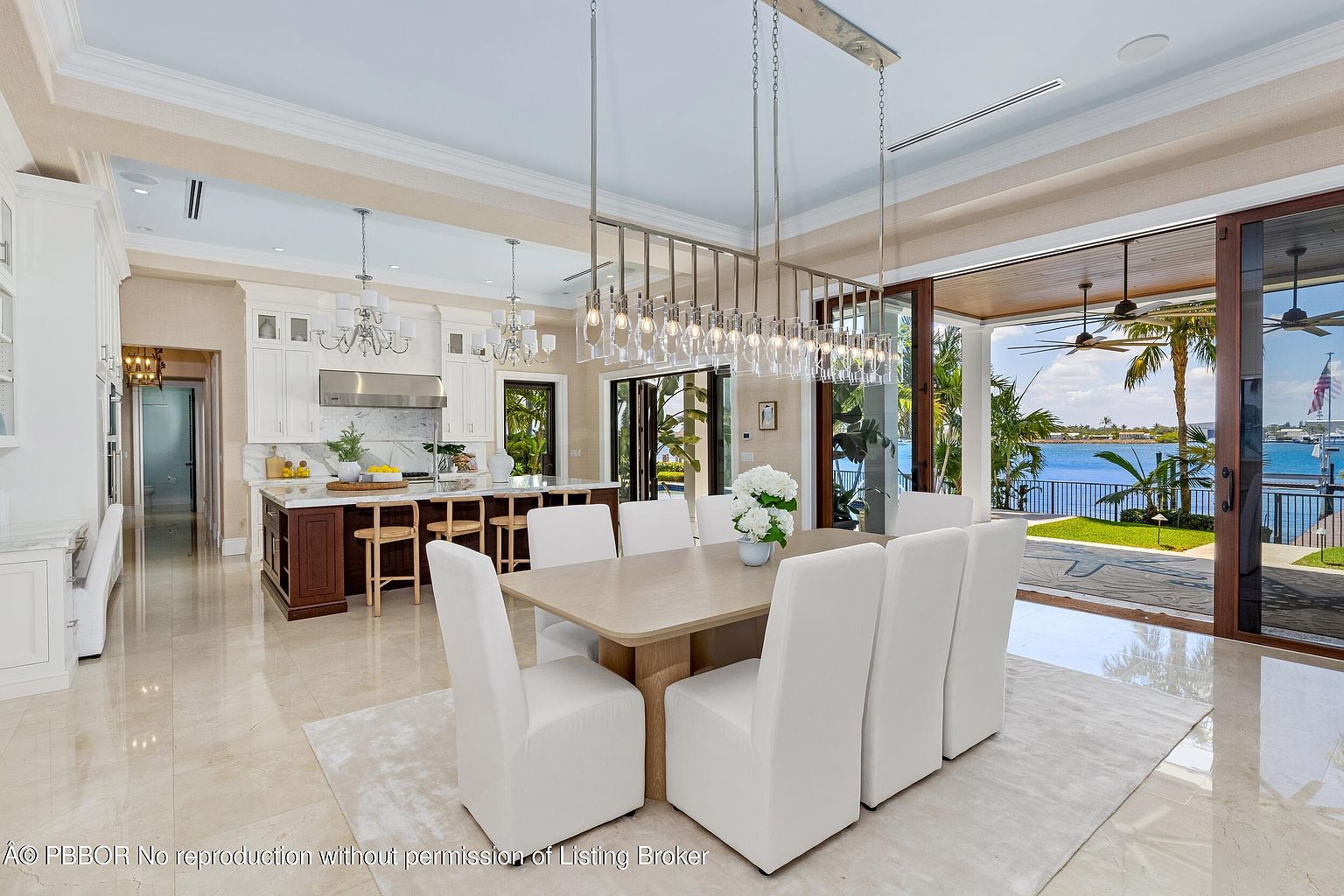 This interior shot showcases a luxurious dining area adjacent to an open-concept kitchen. The dining room features a rectangular table with white upholstered chairs, illuminated by a modern chandelier. Large sliding glass doors offer stunning waterfront views, enhancing the room's appeal and creating a seamless indoor-outdoor living experience.