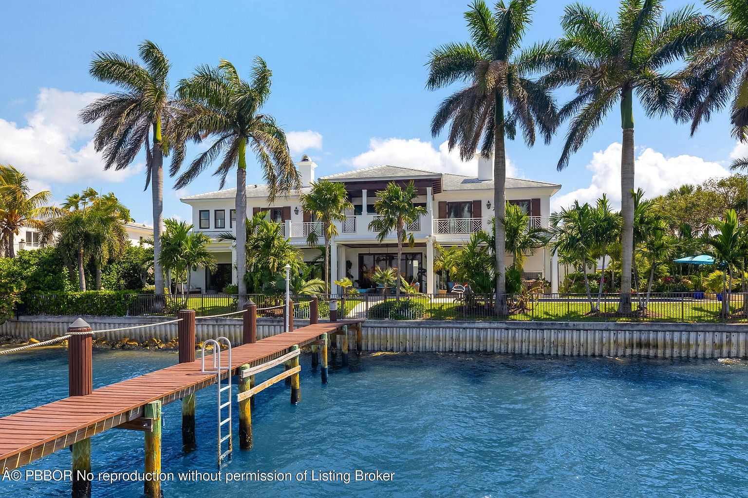 This image showcases the rear exterior of a luxurious waterfront property. The house features a multi-level design with balconies, white facade, and dark shutters, complemented by lush tropical landscaping including mature palm trees. A private wooden dock extends into the clear blue water, enhancing the property's appeal for waterfront living.