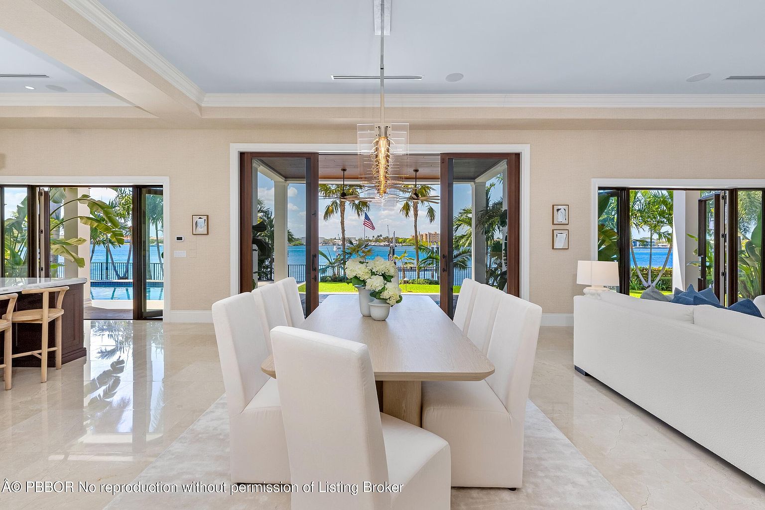 This is an interior shot of a dining room featuring a long, light-wood dining table surrounded by white upholstered chairs. A modern chandelier hangs above the table, and large sliding glass doors offer a view of the waterfront and palm trees. The room is bright and airy, with light-colored walls and flooring, creating a luxurious and inviting atmosphere.