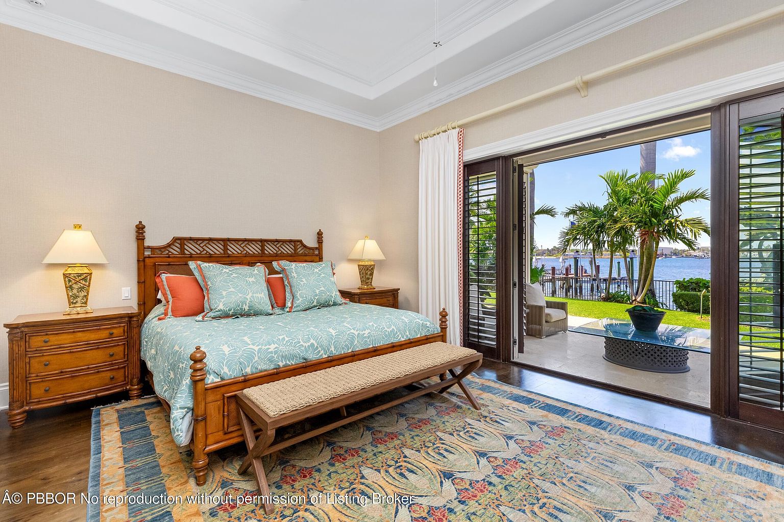 This primary bedroom features a wooden bed frame with patterned bedding and decorative pillows, complemented by matching nightstands and lamps. A woven bench sits at the foot of the bed, and a colorful rug adds warmth to the hardwood floors. Sliding glass doors open to a patio with a view of the water and palm trees, creating a serene and luxurious atmosphere.