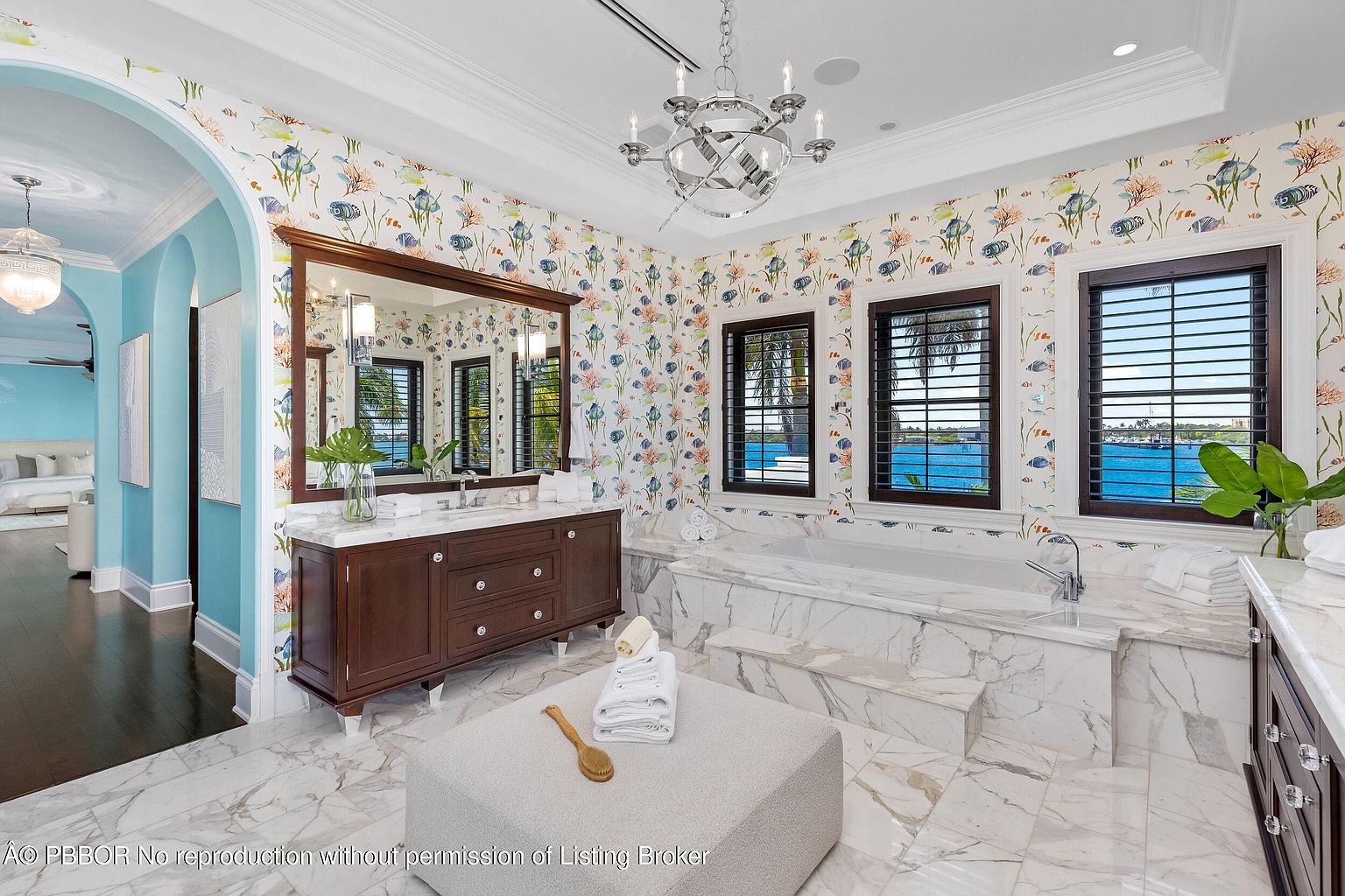 This is a luxurious primary bathroom featuring a marble-clad bathtub and flooring, a dark wood vanity with a marble countertop, and walls adorned with a whimsical fish-themed wallpaper. Natural light floods the space through two windows with dark wood frames, offering a view of the water. A modern chandelier hangs from the ceiling, adding a touch of elegance.
