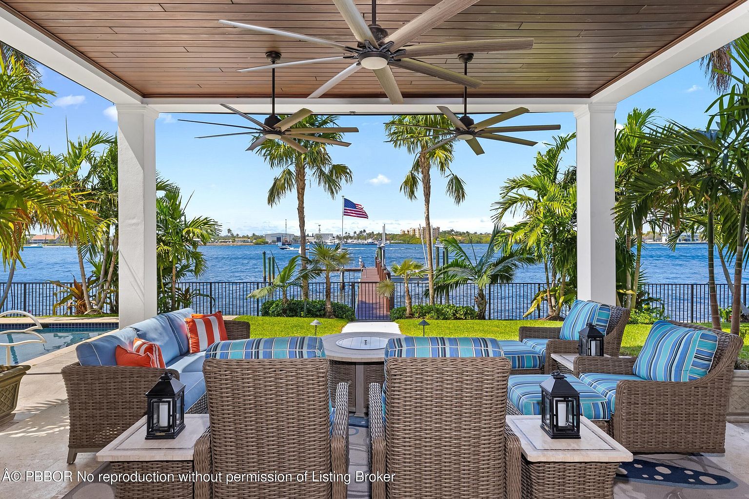 This image showcases a luxurious outdoor patio area with comfortable wicker furniture, including a sofa, chairs, and a central fire pit table. The patio is covered with a wooden ceiling and features ceiling fans, offering a shaded and breezy space. The view extends to a waterfront with palm trees, a dock, and an American flag, creating a serene and inviting atmosphere.