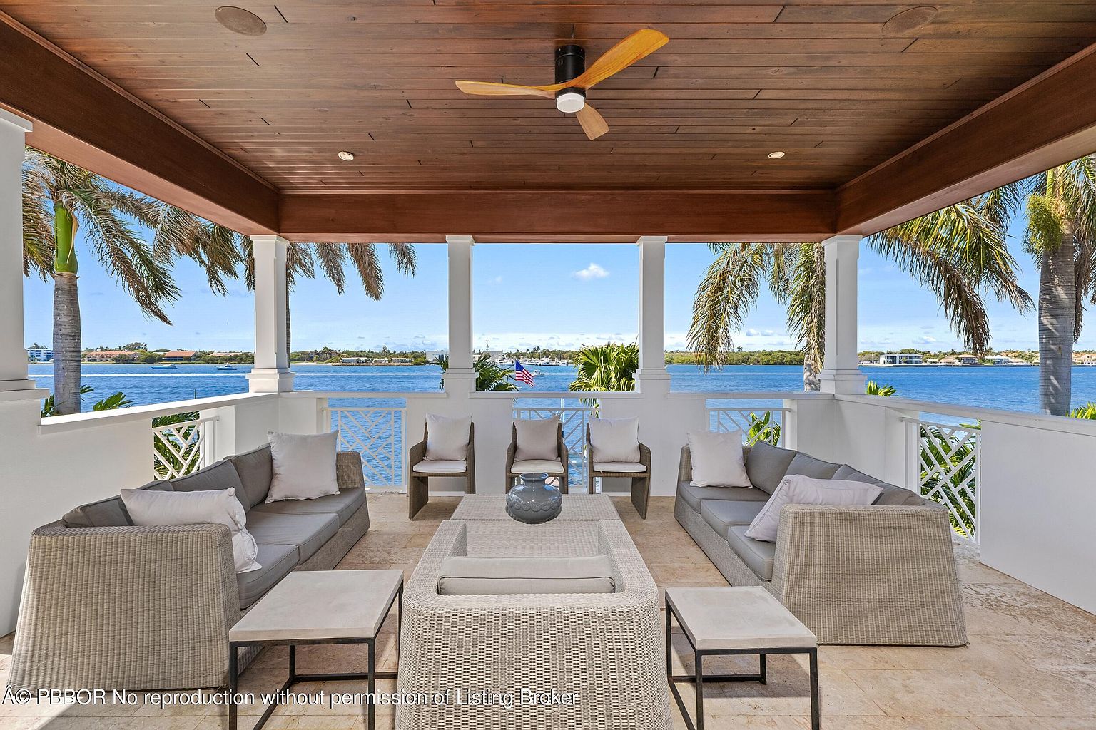 This is a luxurious outdoor patio or balcony area with comfortable seating arrangements, including sofas and chairs, all designed in a neutral color palette. The space is covered by a wooden ceiling with a ceiling fan, and white columns frame a stunning waterfront view. The overall impression is one of relaxation and upscale outdoor living.
