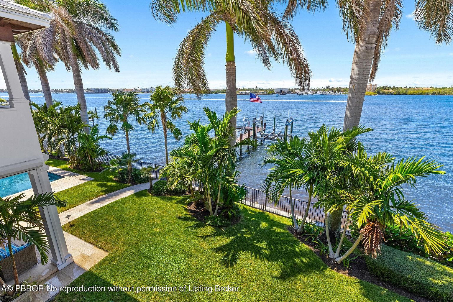 This exterior shot showcases the rear of a luxurious waterfront property. The manicured lawn leads to a private dock with an American flag, set against a backdrop of serene blue water. Palm trees frame the view, enhancing the tropical ambiance and highlighting the property's prime location.