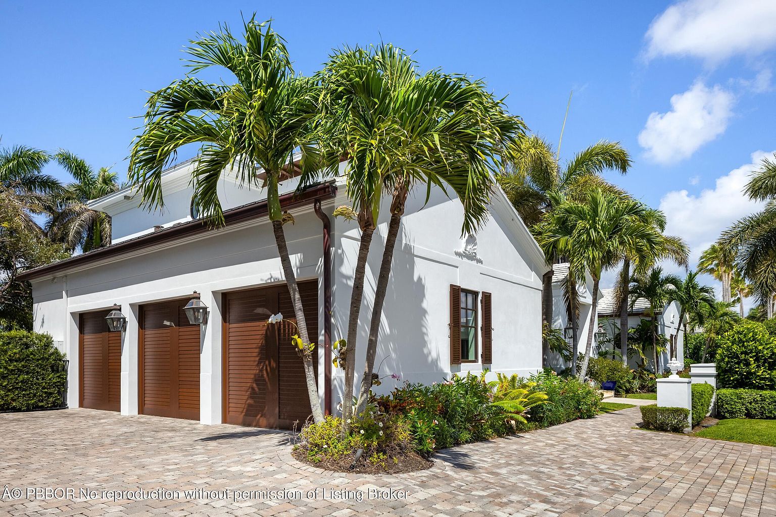 This image showcases the exterior of a home, specifically the garage area. The garage features three wooden doors, complemented by the white exterior walls of the house. Palm trees and manicured landscaping add to the property's curb appeal, while a brick driveway leads up to the garage, creating a welcoming and upscale impression.