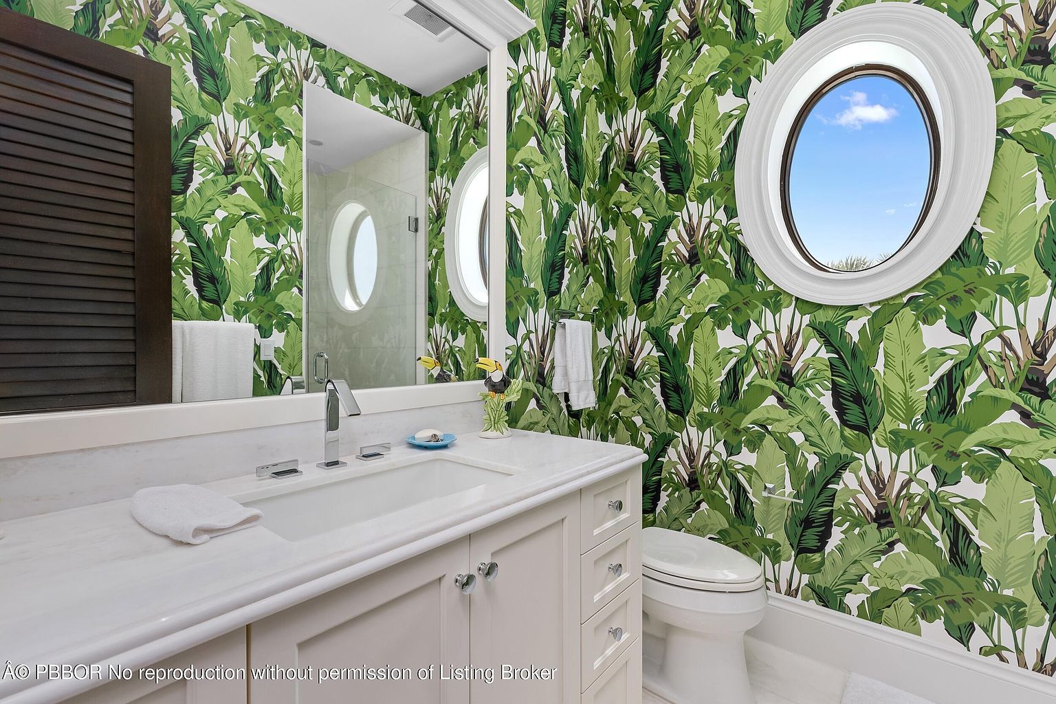 This is a well-lit bathroom featuring a white vanity with a marble countertop, a rectangular sink, and chrome fixtures. The walls are adorned with a vibrant green tropical leaf wallpaper, complemented by a round window that offers a glimpse of the sky. A white toilet is positioned next to the vanity, and the overall impression is fresh and stylish.