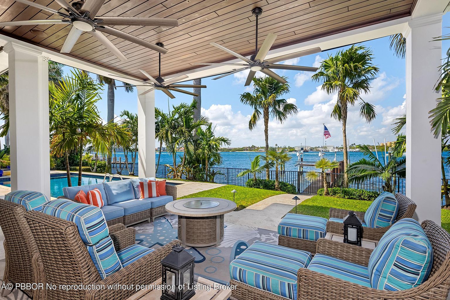 This is a luxurious outdoor patio area with comfortable seating arranged around a central fire pit. The space is covered by a wooden ceiling with ceiling fans, offering shade and ventilation. The patio overlooks a beautiful waterfront view with palm trees and a glimpse of a swimming pool, creating an inviting and relaxing atmosphere.