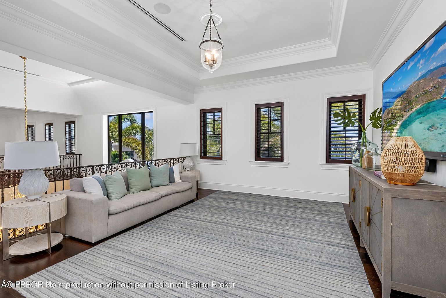 This is an interior shot of a living room area, likely on an upper floor, featuring a neutral-toned sofa with decorative pillows, a light-colored area rug with subtle stripes, and a media console with a large television. The room has a bright and airy feel, with natural light coming through the windows, complemented by a decorative pendant light fixture and recessed lighting.