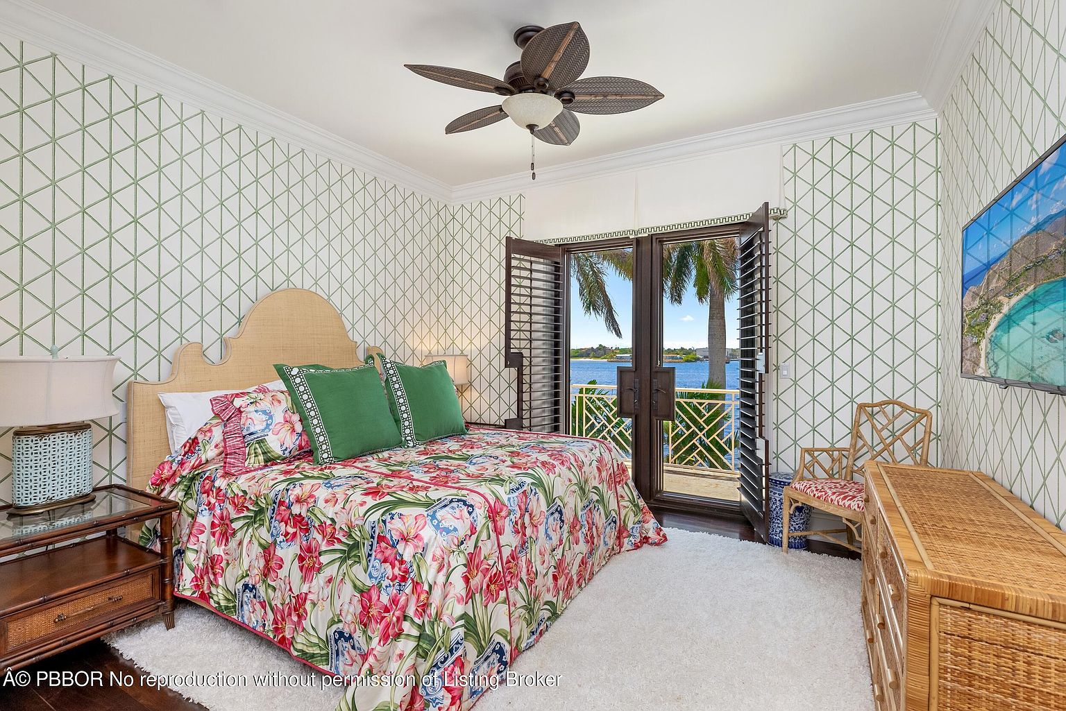 This primary bedroom features a tropical aesthetic with a floral patterned bedspread, green accent pillows, and a woven headboard. The room is bright, with natural light streaming in from the balcony doors, offering a view of the water. A ceiling fan and rattan furniture add to the relaxed, coastal vibe.