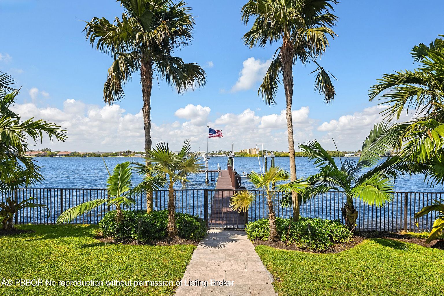 This exterior rear view showcases a waterfront property with a private dock extending into the water, marked by an American flag. The property is framed by lush landscaping, including palm trees and manicured lawns, with a decorative fence and gate leading to the dock. The scene is bathed in sunlight under a partly cloudy sky, creating an inviting and serene atmosphere.