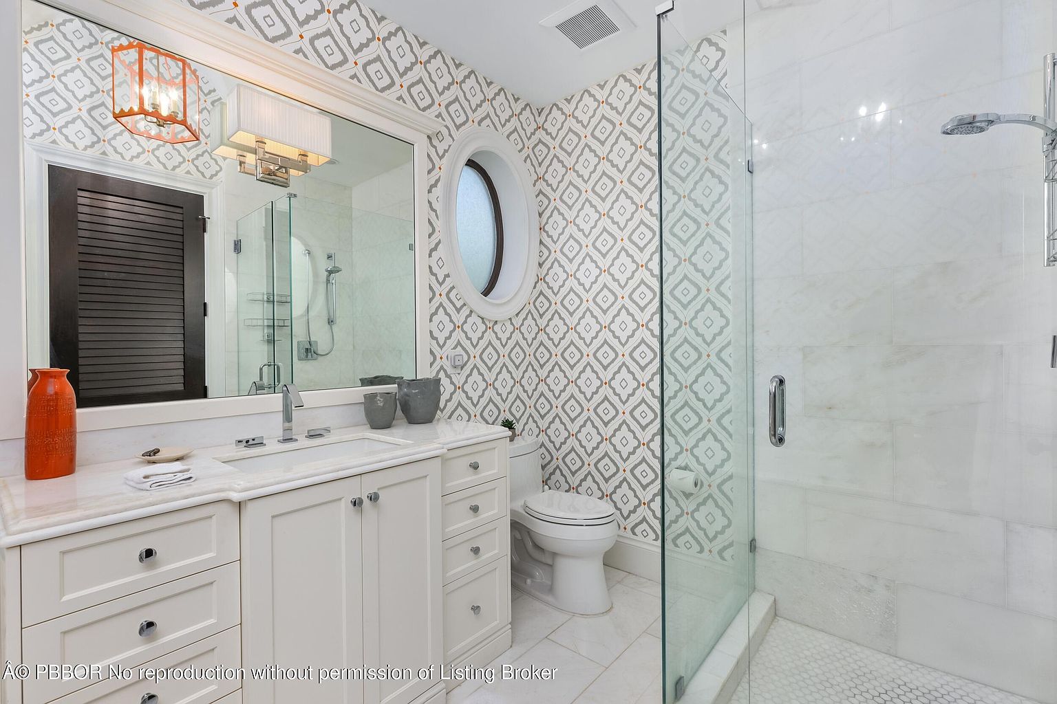 This is a well-lit primary bathroom featuring a white vanity with marble countertop, a large mirror, and patterned wallpaper. A glass-enclosed shower is visible to the right, and an oval window adds a unique architectural detail. The overall impression is clean, elegant, and modern.
