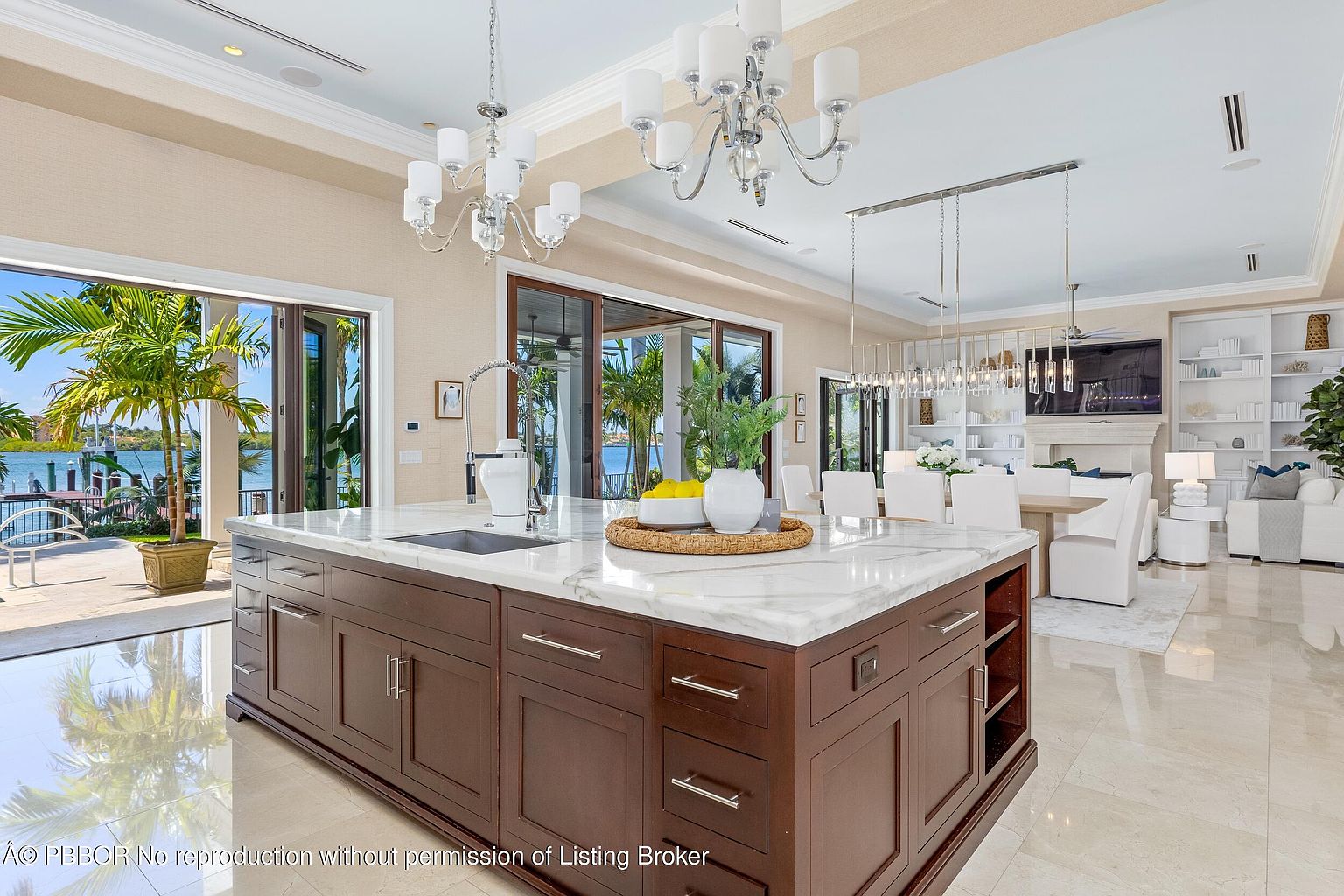 This is a bright and luxurious kitchen featuring a large center island with a marble countertop and dark wood cabinetry. The space is illuminated by elegant chandeliers, and large sliding glass doors offer views of the waterfront. The open floor plan seamlessly connects the kitchen to the dining and living areas, creating an inviting and sophisticated atmosphere.