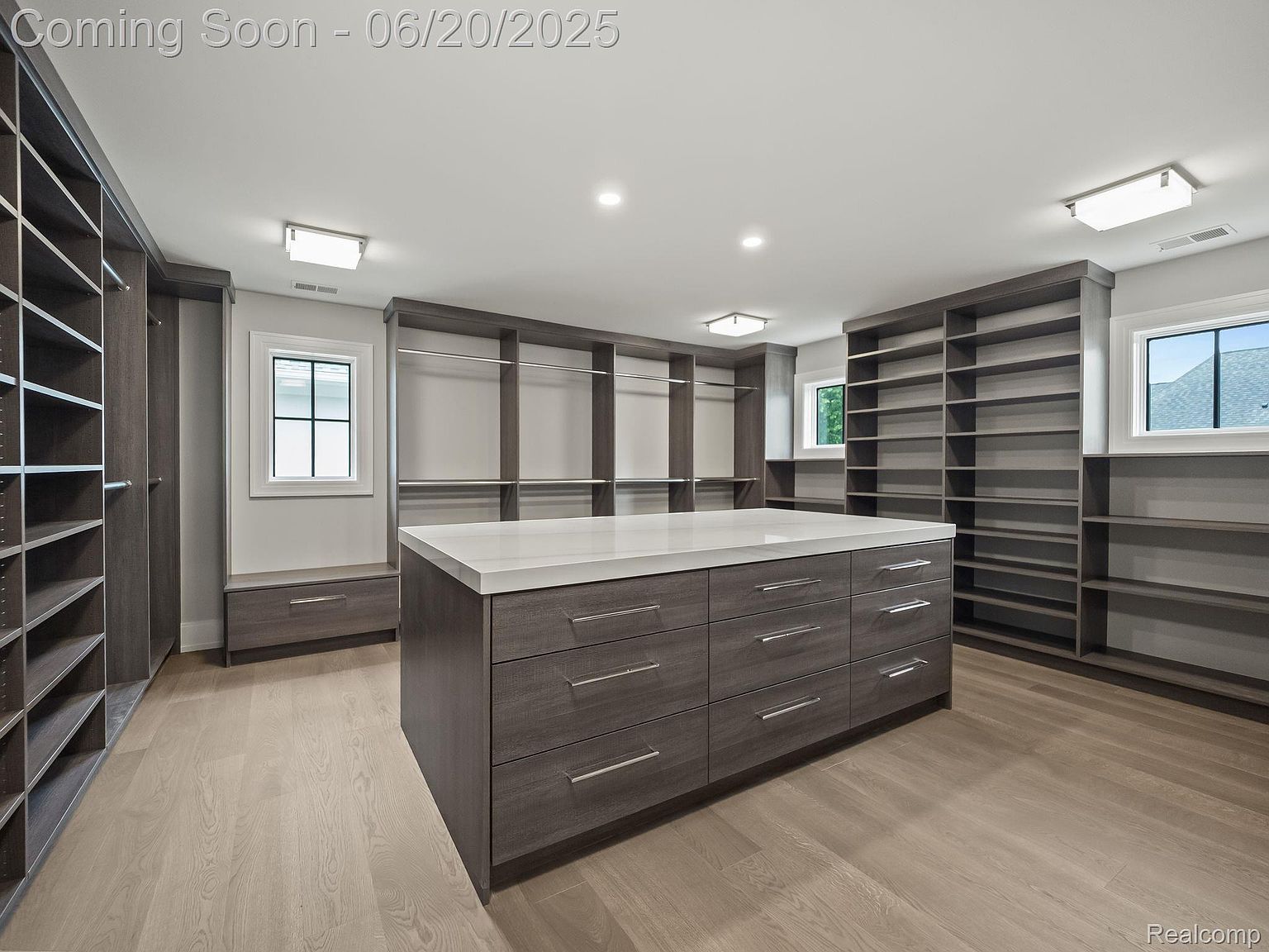This is a spacious walk-in closet featuring custom shelving and drawer units in a dark wood finish. A large center island with a white countertop provides ample space for organizing and displaying items. The closet is well-lit with recessed lighting and natural light from the windows, creating a luxurious and functional space.