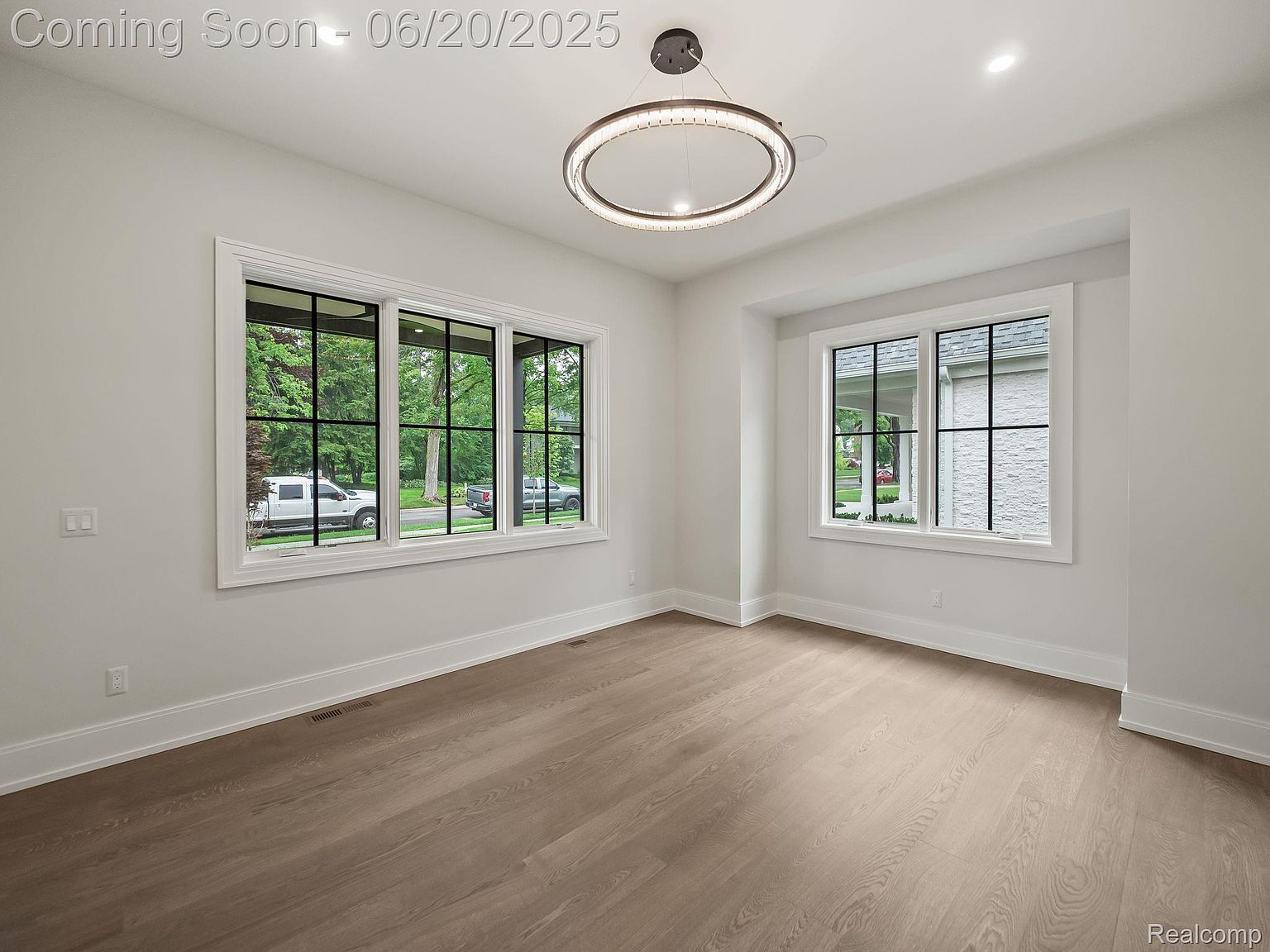 This is an interior shot of a dining room featuring hardwood floors, white walls, and large windows that provide ample natural light. A modern, circular chandelier hangs from the ceiling, adding a touch of elegance. The room is spacious and appears to be newly constructed or renovated, giving a clean and contemporary feel.