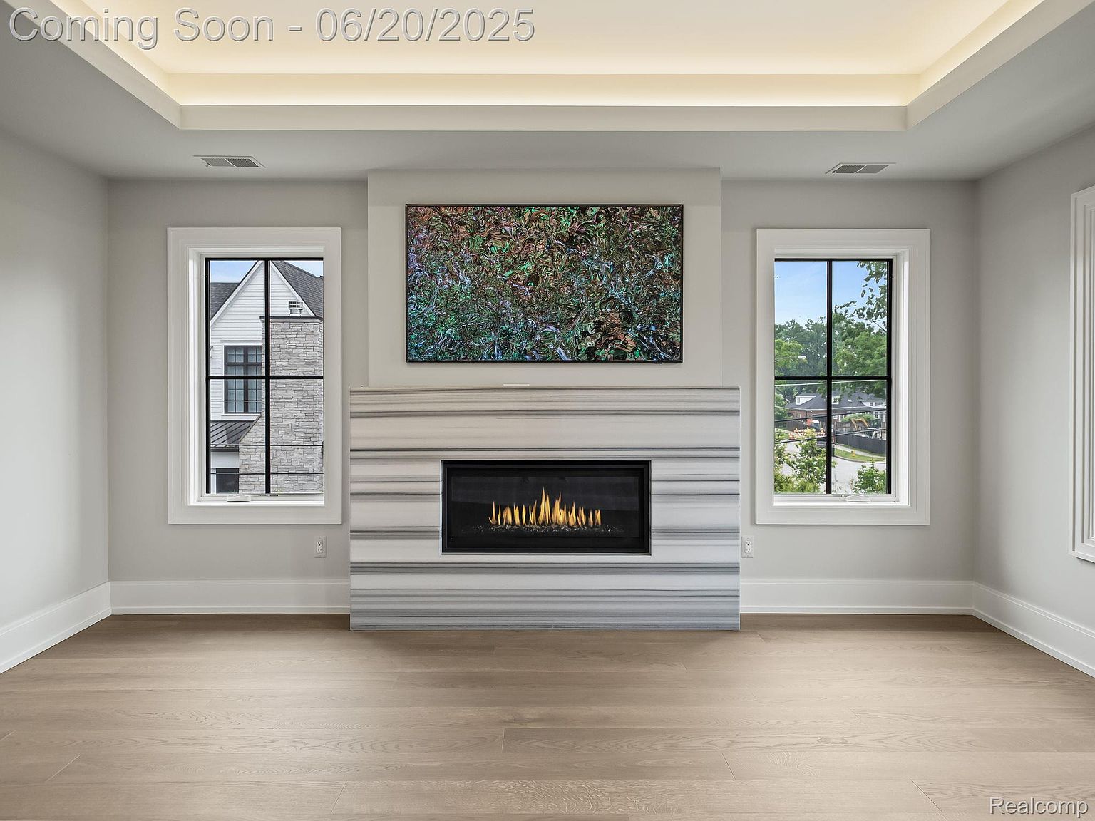 This is an interior shot of a living room featuring a modern fireplace with a marble surround and a large abstract painting above it. Two windows flank the fireplace, providing natural light and views of the outside. The room has light hardwood floors and neutral-colored walls, creating a bright and airy atmosphere.