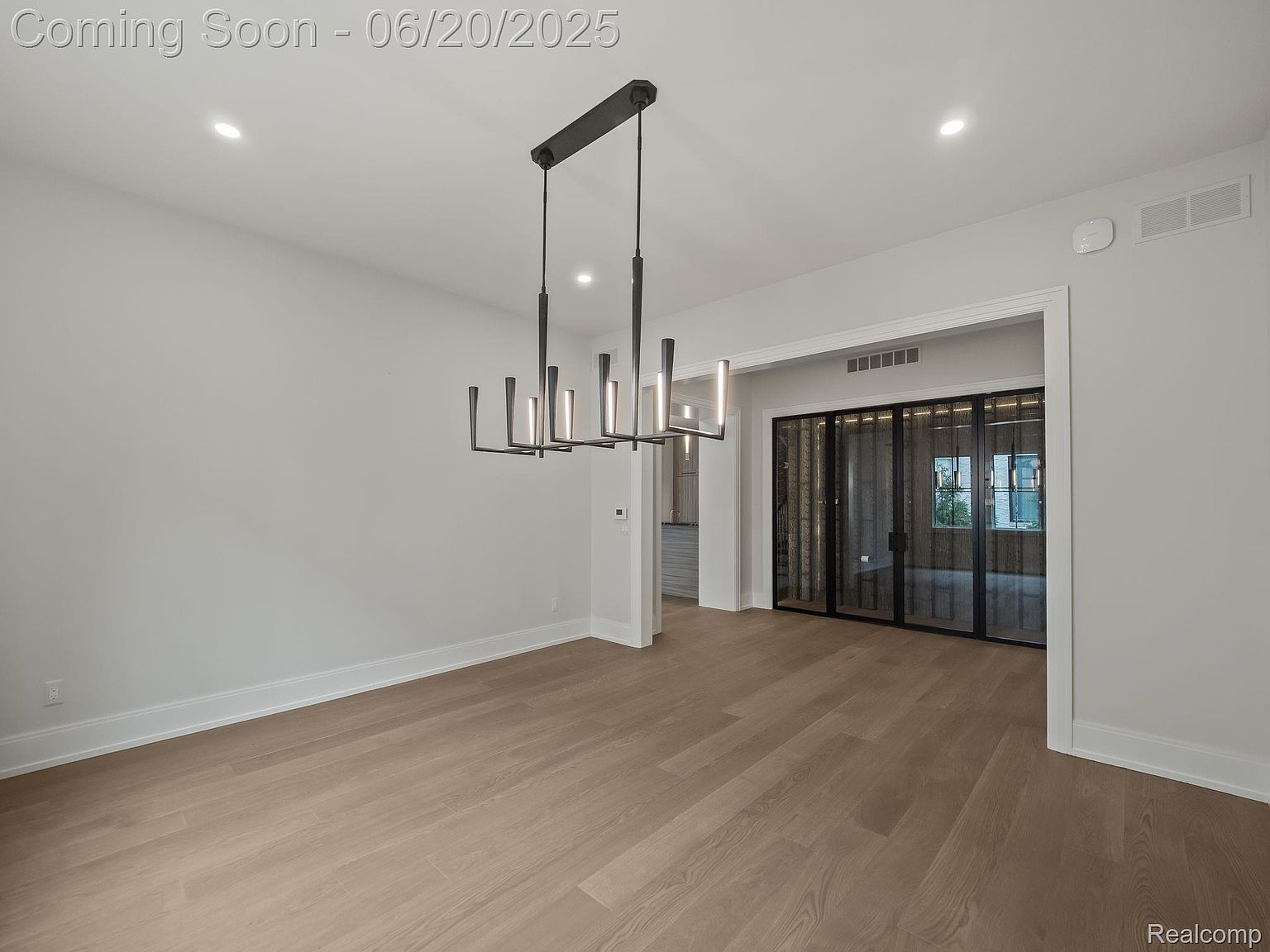 This is an interior shot of a dining room featuring light hardwood floors and neutral gray walls. A modern, linear chandelier hangs above the center of the room, providing a focal point. A doorway leads to another room with sliding glass doors, adding depth and visual interest to the space.