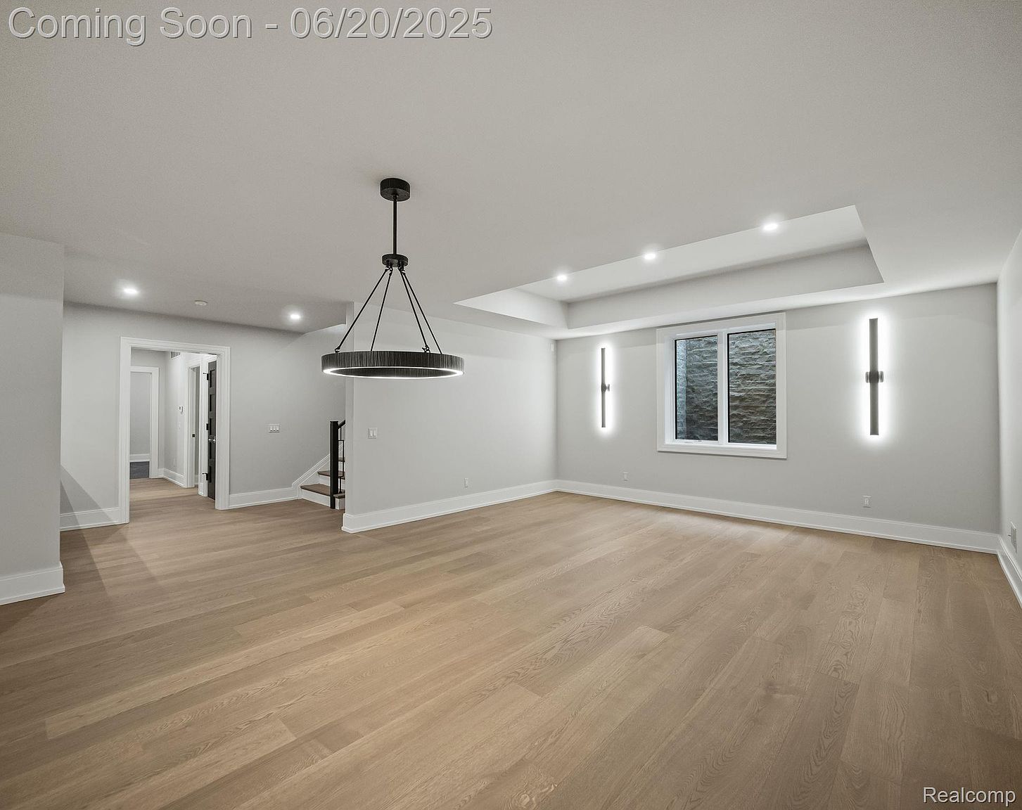 This is a spacious living room featuring light hardwood floors and neutral gray walls. A modern, circular chandelier hangs from the ceiling, and recessed lighting adds to the brightness. A window with a view to the outside is flanked by modern sconces, and an open doorway leads to another room, creating an inviting and airy atmosphere.