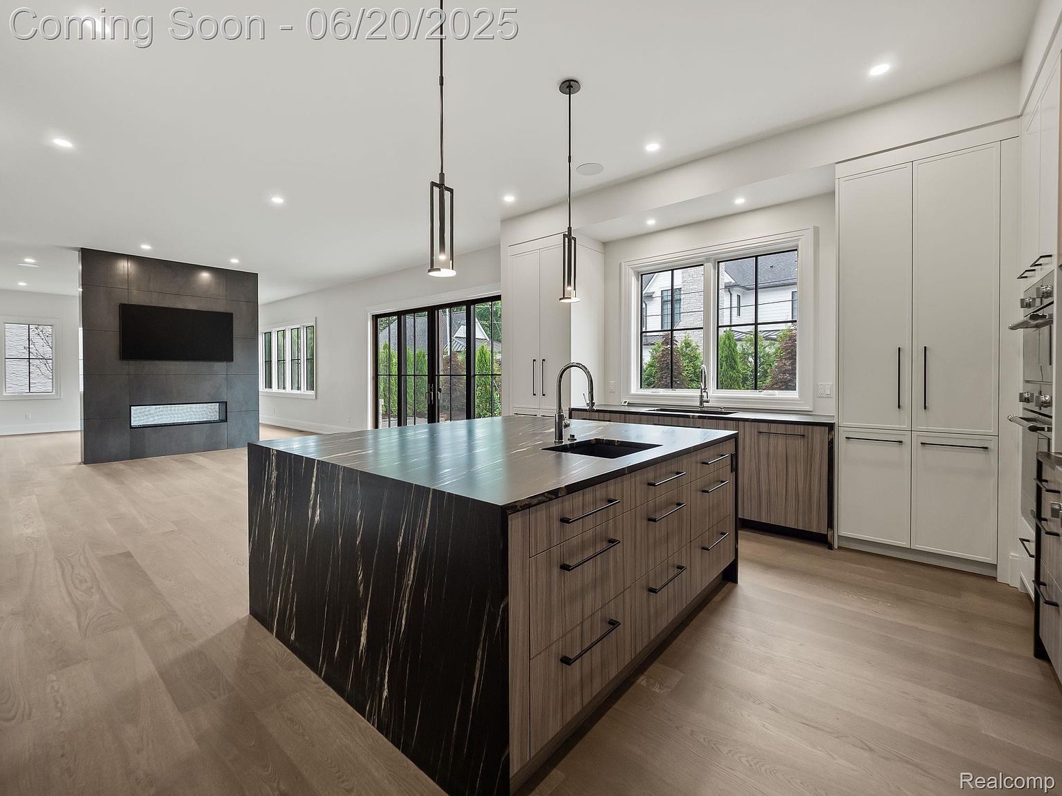 This is a bright and modern kitchen featuring a large island with a dark countertop and wood-grain cabinets. The kitchen includes stainless steel appliances, white cabinetry, and a window overlooking the backyard. The open floor plan connects the kitchen to the living area, which features a fireplace and a large television.