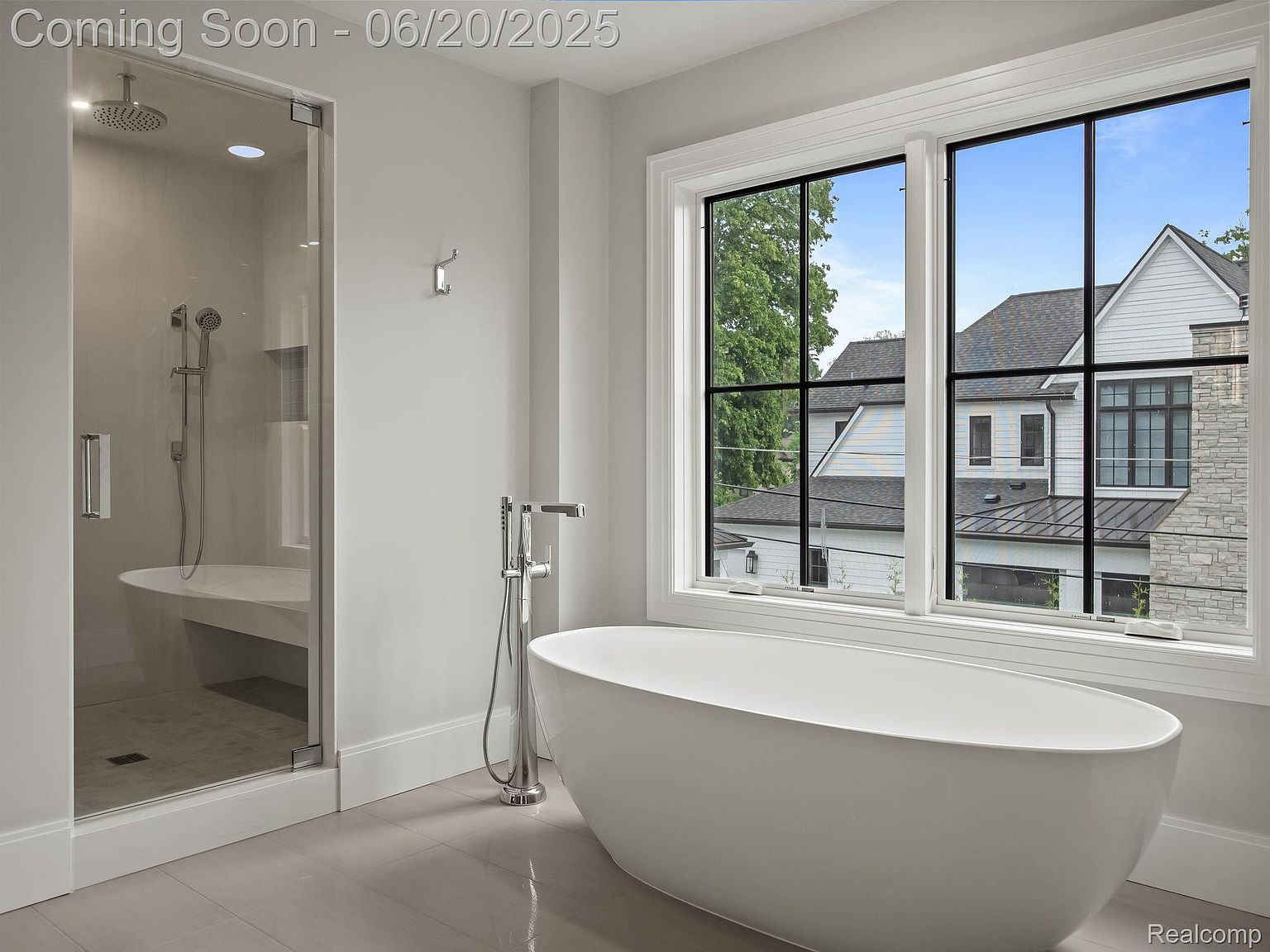 This is a bright and modern primary bathroom featuring a glass-enclosed shower with a built-in bench and a freestanding soaking tub. The room is painted in a neutral color palette, with a large window providing natural light and a view of the exterior. The overall impression is clean, luxurious, and spa-like.
