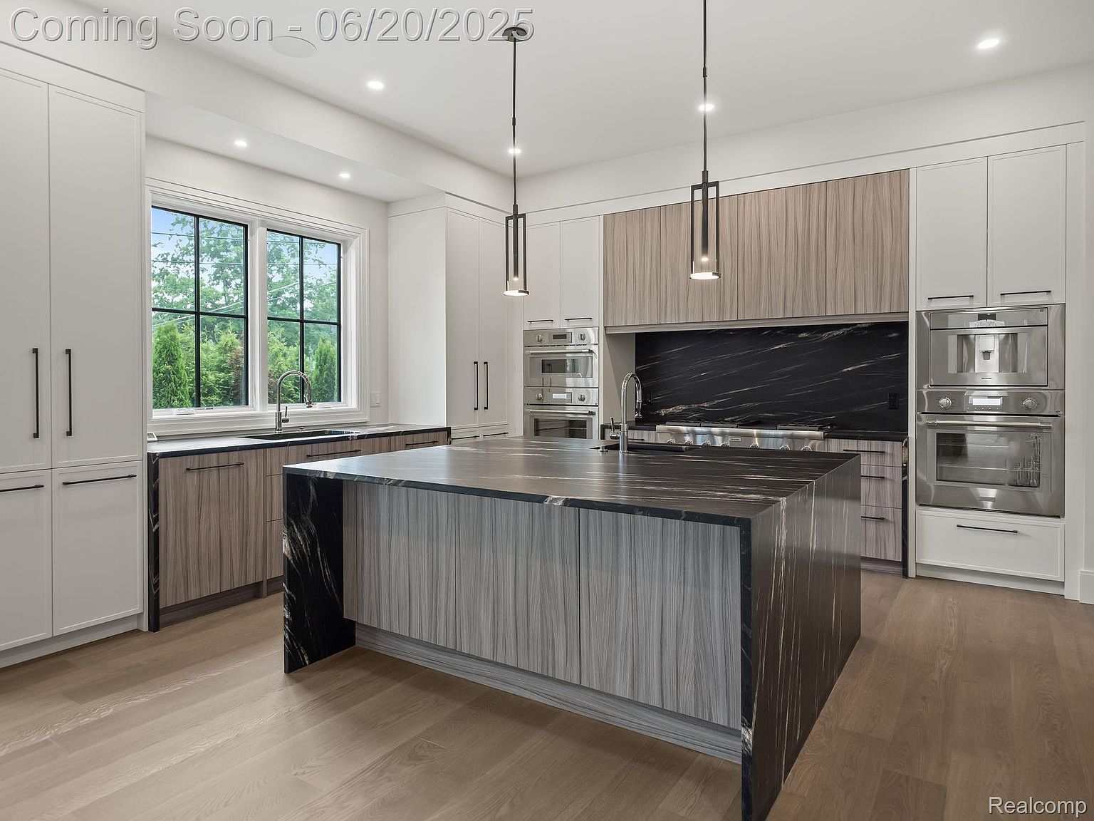 This is a modern kitchen with a large island featuring a waterfall countertop and wood-grain cabinetry. The kitchen also includes stainless steel appliances, a black marble backsplash, and a large window that provides natural light. The overall impression is one of luxury and sophistication.