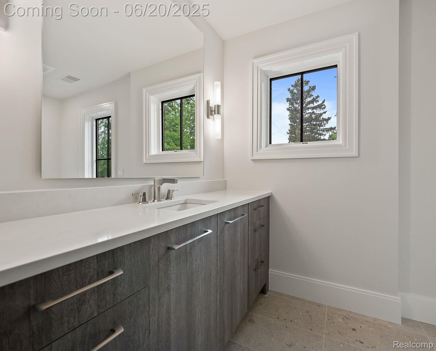 This is a well-lit bathroom featuring a modern vanity with a gray wood-grain finish and a white countertop. A large mirror reflects the window and the vanity, enhancing the sense of space. The room is painted in a neutral color, and the flooring appears to be tile, creating a clean and contemporary aesthetic.