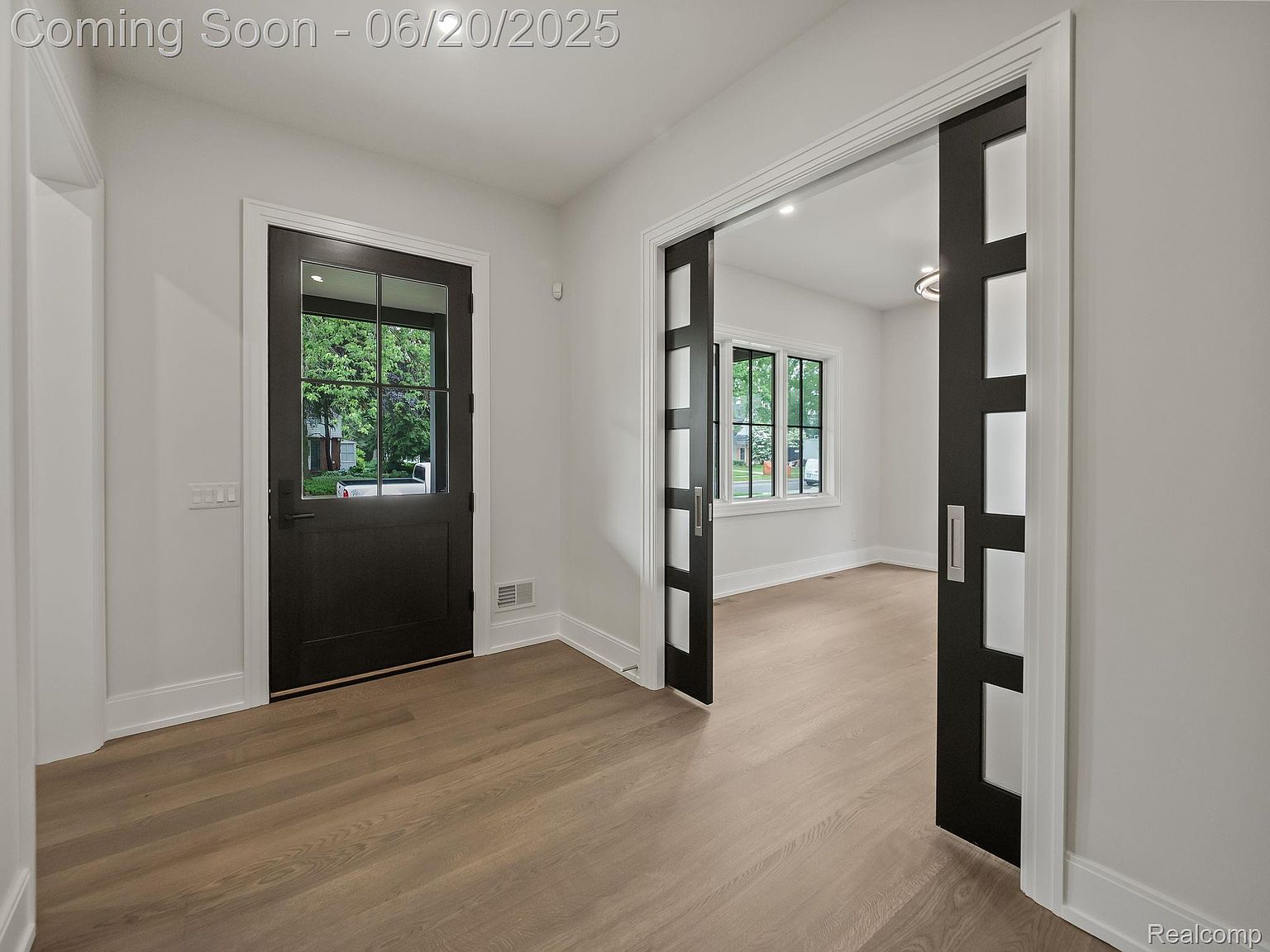 This interior shot showcases a bright hallway with light wood flooring and white walls. A dark-framed glass door leads to the outside, while sliding doors with a modern design open to another room. The space feels clean and inviting, highlighting the home's contemporary style.