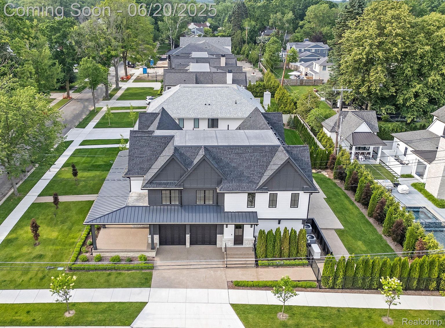 This aerial view showcases a modern, luxury home with a dark gray and white exterior, complemented by a meticulously landscaped yard. The property features a multi-car garage, a well-maintained lawn, and mature trees lining the street, creating a sense of privacy and curb appeal. The overall impression is one of sophistication and high-end living.