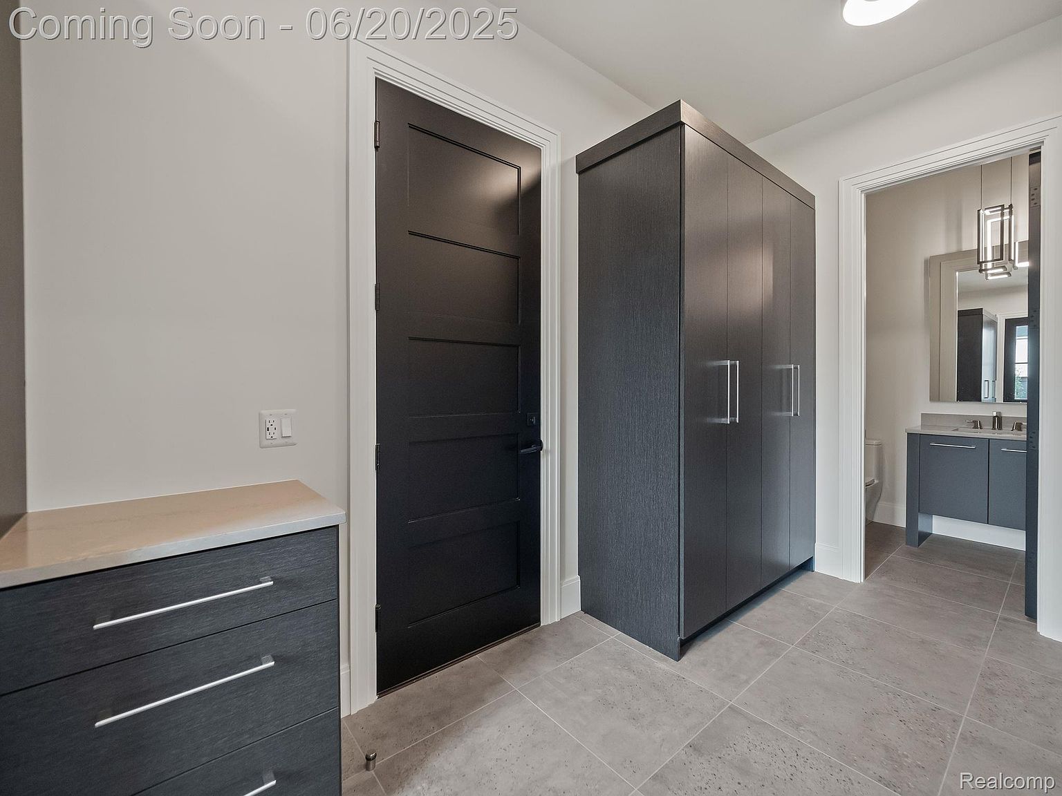 This interior shot showcases a modern hallway featuring a dark-colored door with a white frame, a tall dark-colored storage unit with sleek handles, and a glimpse into a bathroom. The flooring is a light gray tile, and the walls are painted in a neutral tone, creating a clean and contemporary aesthetic.