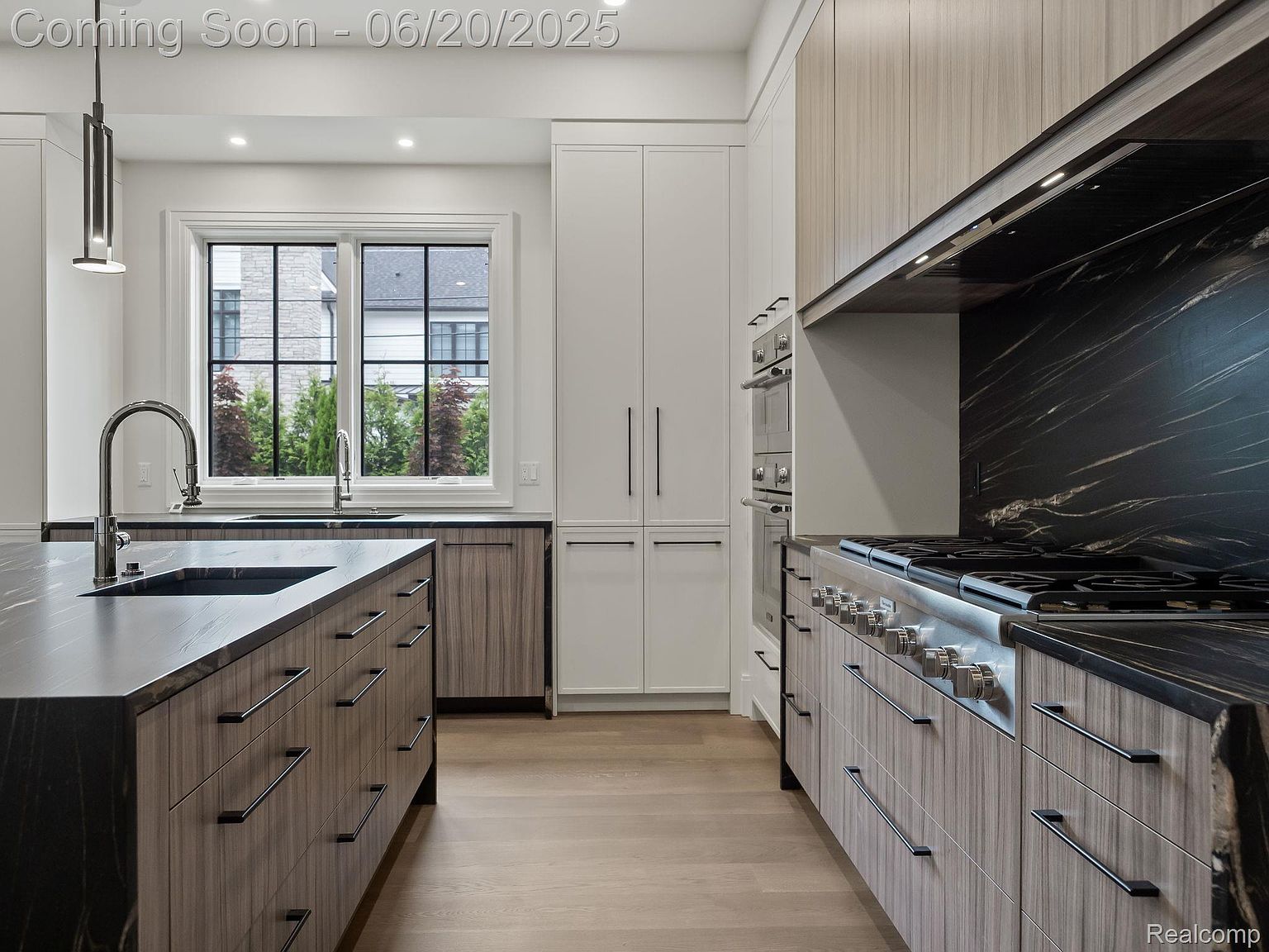 This is a modern kitchen featuring sleek, flat-panel cabinetry in a light wood grain and white, complemented by dark countertops with striking veining. Stainless steel appliances, including a professional-grade range and built-in ovens, add to the high-end feel. The kitchen island provides ample workspace and storage, while a large window offers natural light and a view of the outdoors.