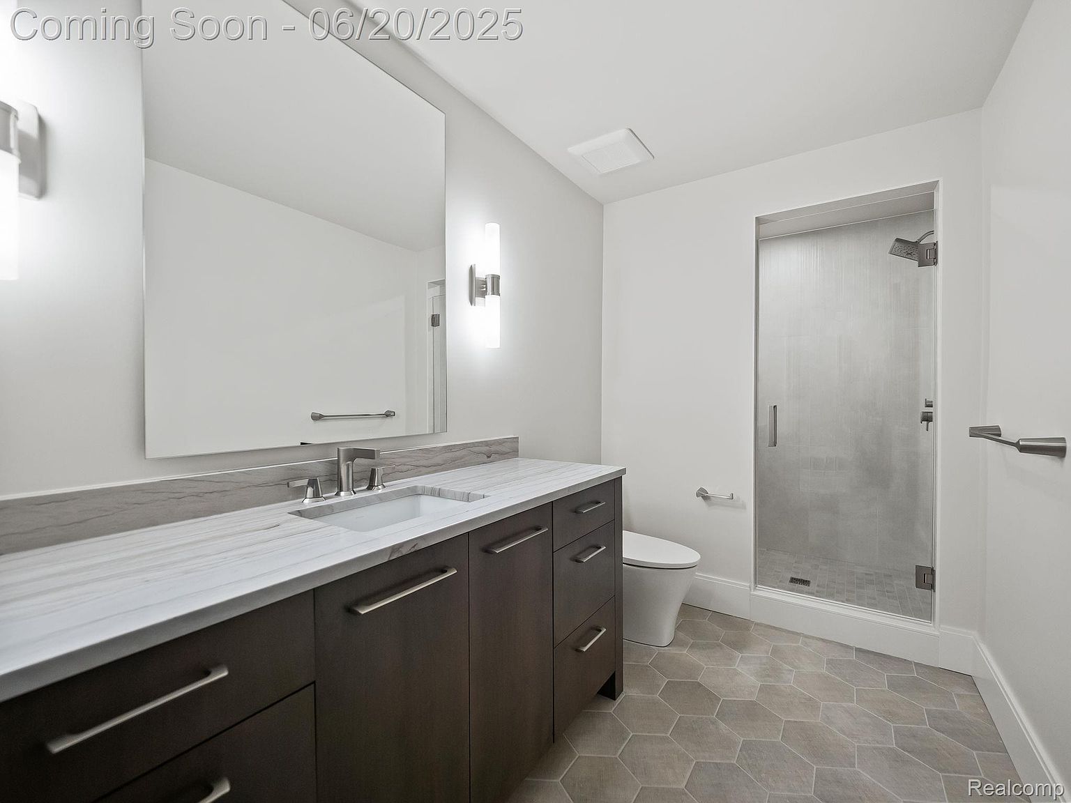 This is a well-lit bathroom featuring a modern vanity with dark wood cabinets and a white countertop, a large mirror, and sleek lighting fixtures. The bathroom also includes a toilet and a glass-enclosed shower with gray tiling. The floor is covered in gray hexagonal tiles, contributing to the contemporary aesthetic.