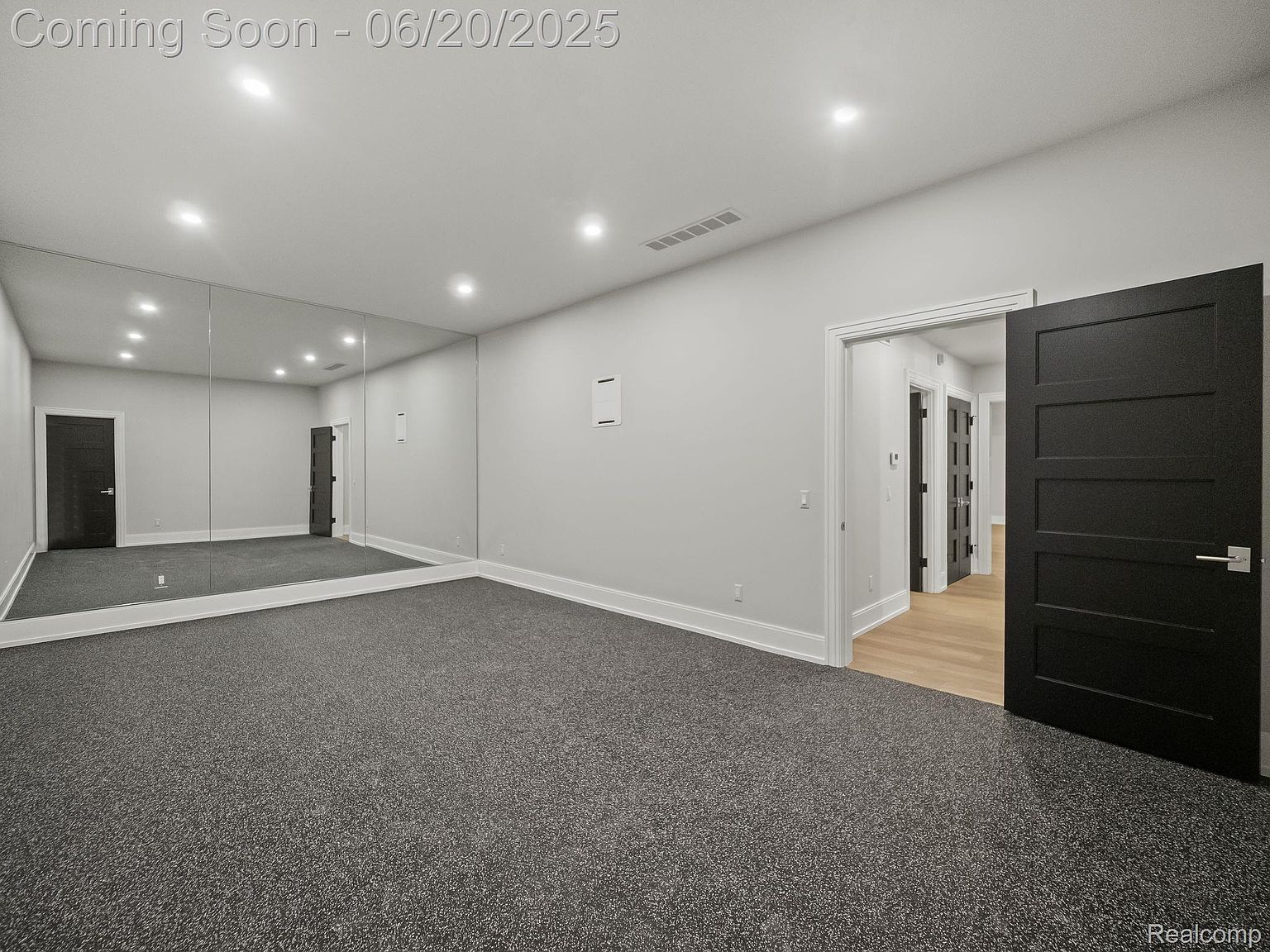 This is an interior shot of a home gym featuring a large mirror wall, gray carpet, and recessed lighting. A dark-colored door is visible on the right, slightly ajar, leading to another room with hardwood flooring. The room appears spacious and well-lit, creating an inviting atmosphere for exercise.